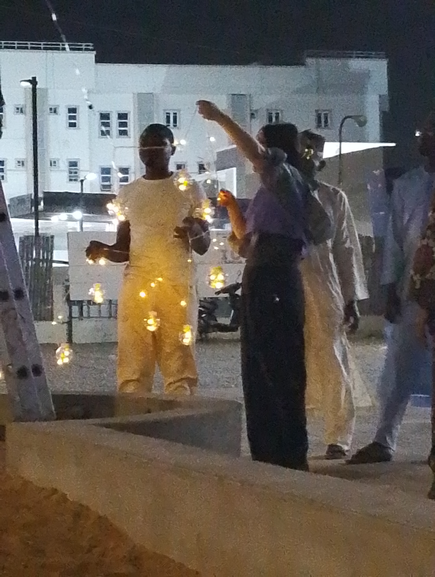 Captured at night in Gandu Albasa, Nigeria, the photograph focuses on a gathering of individuals near a building illuminated by interior lights. One person stands out, wrapped in strings of lights, their outfit and appearance brightly lit. To their right, two men dressed in traditional attire are observed. The scene includes elements of a roadside setting, marked by the presence of a parked motorcycle and a ladder leaning against a pole.