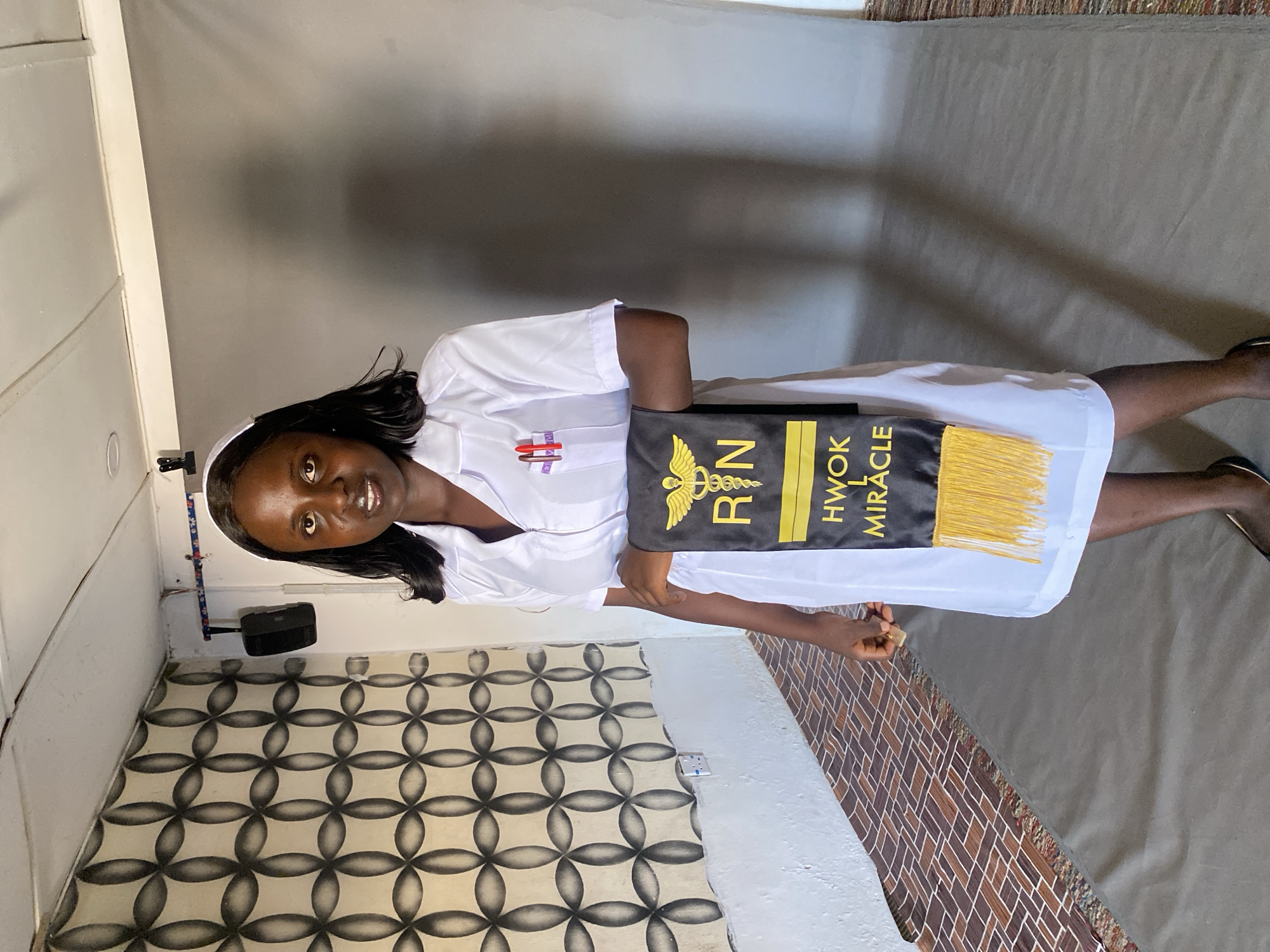 The image shows a young woman in a white nursing uniform and cap, holding a black and gold sash with "RN" and "HWOK MIRACLE" printed on it. She is standing in a room with patterned tile flooring and a gray wall in Vom, Nigeria. A wall-mounted black speaker and an electrical outlet are visible. The floor is decorated with a brown carpet with a brick-like pattern.