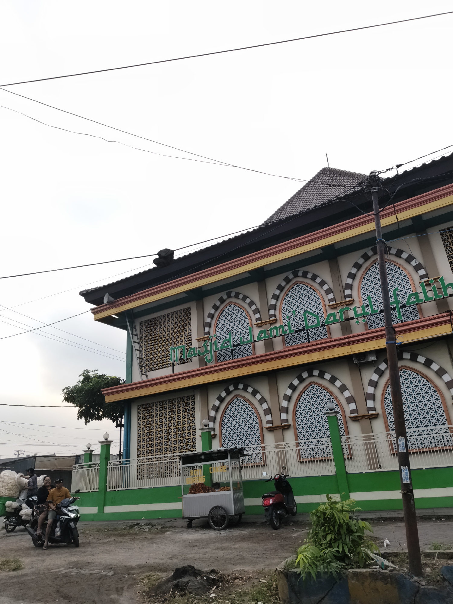 The image depicts a street scene in Sidorejo, Indonesia. The focal point is a multi-story building, likely a mosque, with green and yellow accents and arched windows featuring intricate patterns. Green fencing runs along the front of the building, with a street in front. A food cart is parked to the right of the building. Two people are riding a motorcycle on the street, and another motorcycle is parked nearby. To the left of the scene, a person is seen on a motorcycle carrying a large load of what appears to be white, fluffy material. Power lines crisscross the sky above. The ground is a dirt or gravel surface.