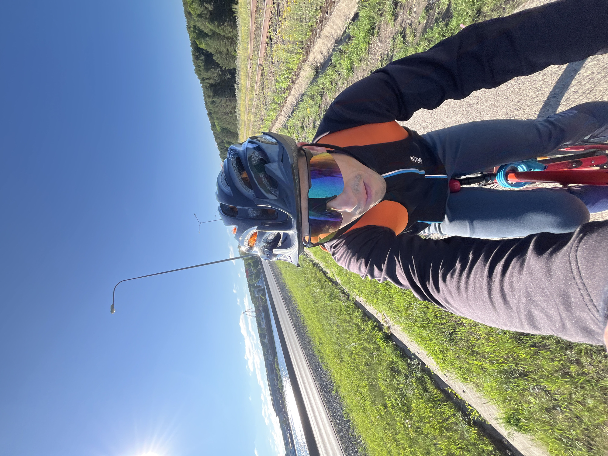 A first-person perspective captures a cyclist wearing a black helmet and reflective goggles. The cyclist is on a paved path next to a body of water, with a road and a forested hill in the background. The sky is clear and blue with a bright sun. The location is the embankment of the Kama River in the city of Chaikovsky, Russia. The path appears to be a dedicated cycling route with a grassy verge. In the distance, industrial structures and a roller coaster-like amusement ride are visible across the river. The cyclist is wearing a black long-sleeved jacket with orange accents and grey pants.