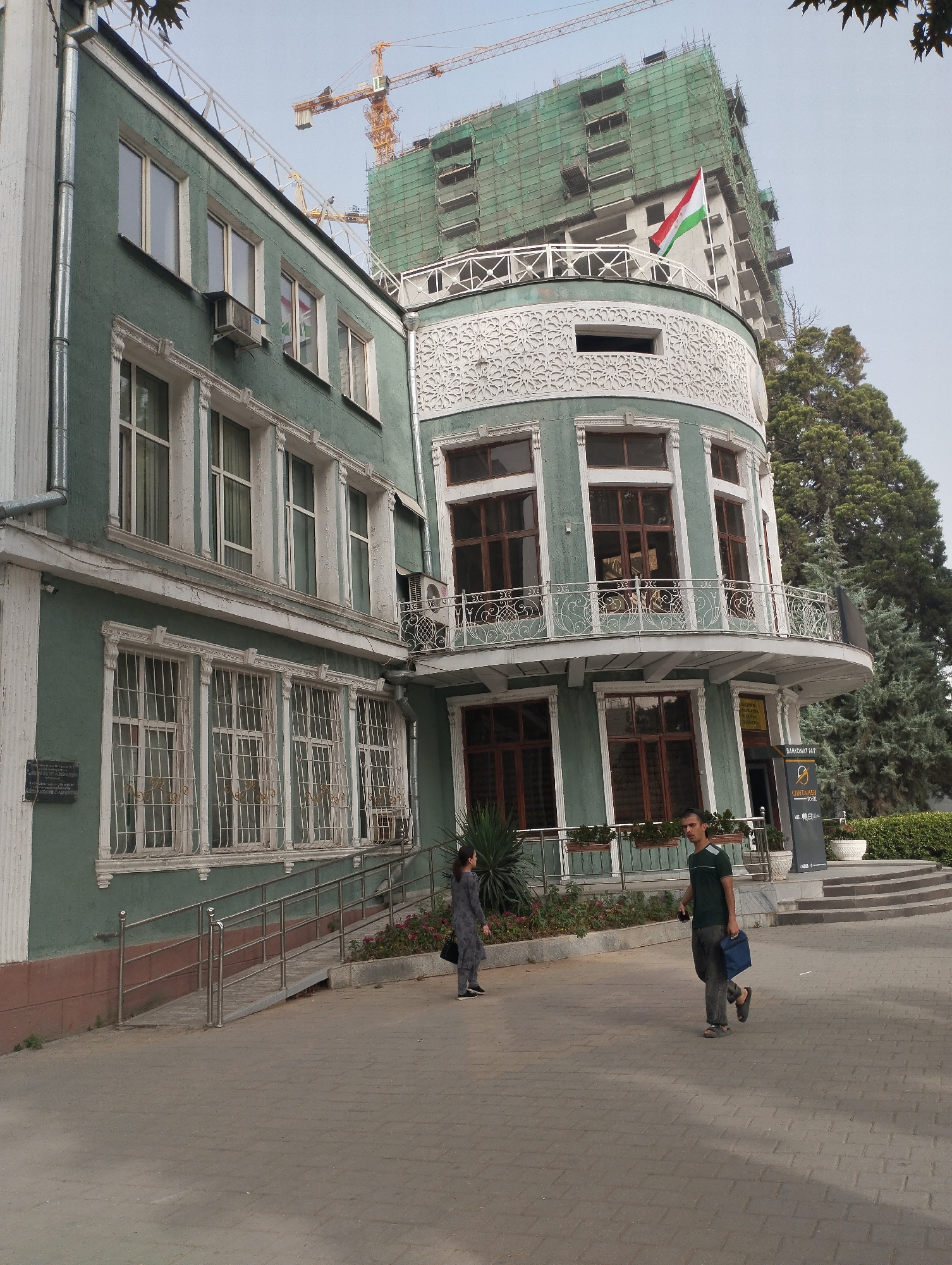 The image depicts a street-level view of a building in Dushanbe, Tajikistan, likely near the National Museum of Tajikistan. The building itself is an older, pastel green structure with ornate white trim around its windows and a rounded, multi-story section in the foreground. Above and behind this structure, a tall, modern building is under construction, covered in green netting and topped with a scaffolding. A flag, appearing to be the flag of Tajikistan (red, white, and green horizontal stripes), is visible on the construction site.
In the foreground, a paved walkway leads towards the building. Two individuals are walking on the walkway. To the left, a woman in a dark grey patterned outfit is walking towards the building entrance, carrying a black bag. To the right, a man in a dark green t-shirt and dark pants is walking away from the building, carrying a blue bag. He is looking towards the left. The walkway is bordered by planters containing greenery and flowers, and a ramp with metal railings leads up to the building's entrance. In the background, a large evergreen tree stands to the right of the buildings. The sky is overcast. A crane is visible in the far background, indicating ongoing construction activities in the vicinity.