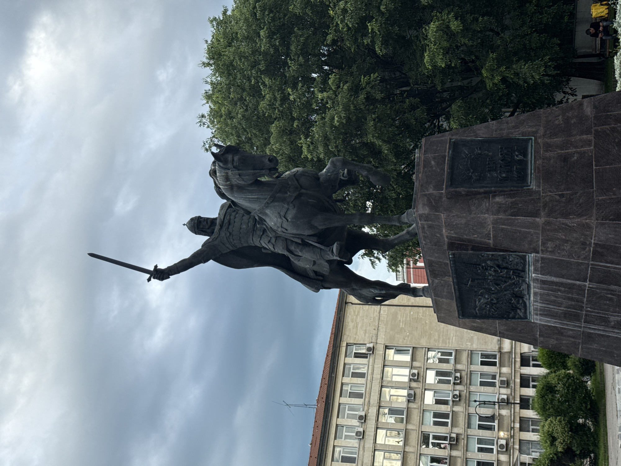 The image shows a bronze statue of a warrior on horseback holding a sword aloft, likely a historical figure. The statue is mounted on a dark, stone plinth with bronze plaques affixed to its sides. Behind the statue is a grey, overcast sky and a large green tree. A multi-story building with many windows, most of which appear to have air conditioning units, is also visible in the background. The image location is likely in Varna, Bulgaria, based on the architectural style of the building and the design of the monument. There appear to be at least two people sitting in the upper right.