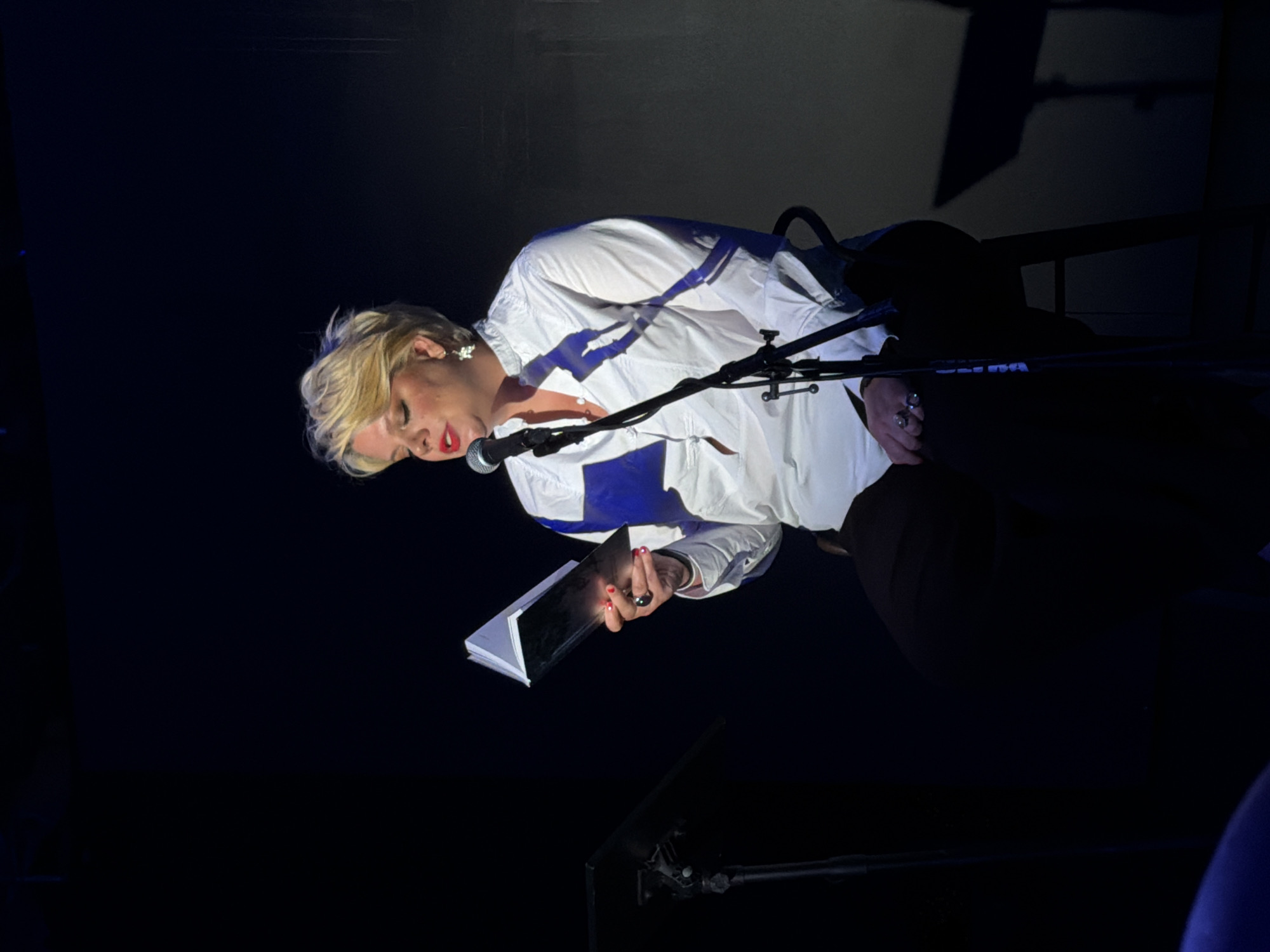 In a dimly lit indoor venue, a woman with short blonde hair and red lipstick is seated on a stool and reading from a book. She is wearing a white collared shirt with buttons and dark pants, and she has rings on her fingers. A microphone on a stand is positioned in front of her. To her left, a music stand is visible. The background is dark, with some indistinct shapes and lighting suggesting a stage or performance area. The overall impression is of a performance or reading. The specific location in Los Angeles, California, USA, cannot be determined from the image alone.