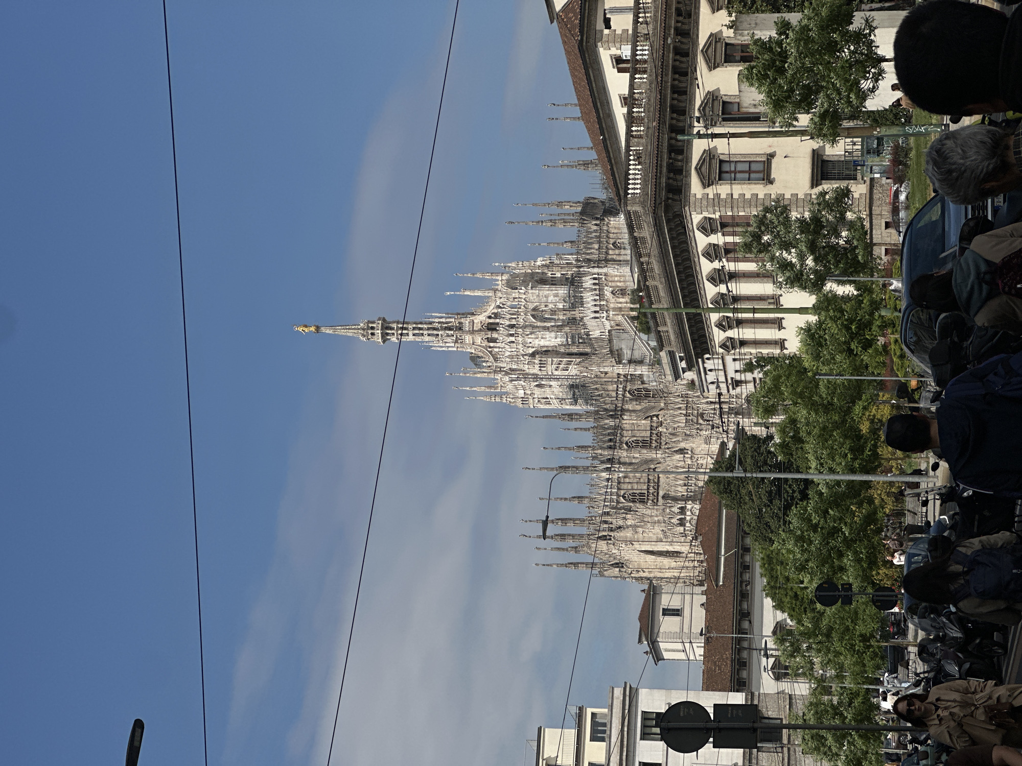The image depicts the Milan Cathedral, or Duomo di Milano, in Milan, Italy, viewed from a street-level perspective. The cathedral's façade, with its intricate spires and sculptures, is partially obscured by utility lines against a blue sky with wispy clouds. Adjacent to the cathedral is a building with a detailed façade. The foreground includes trees, street signs, parked motorcycles, and pedestrians. People are seen in groups, suggesting a tourist area. The day is bright, with natural light illuminating the scene.