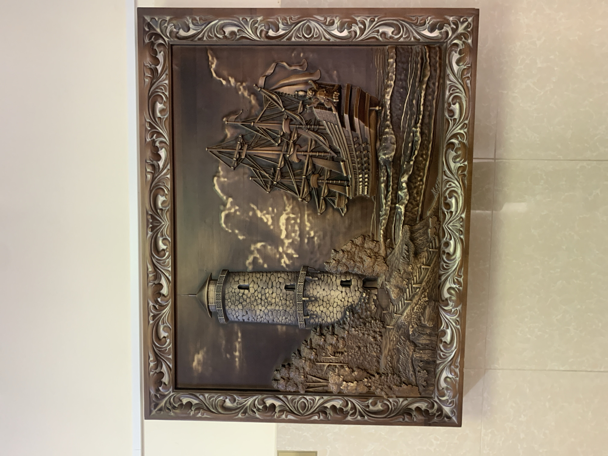 This is a photograph of a framed bas-relief artwork depicting a maritime scene. The artwork is mounted on a white wall, with a tiled wall visible on the right. The artwork itself is rectangular and oriented vertically.
The primary subjects are a tall sailing ship and a stone lighthouse. The ship, rendered in intricate detail, is shown with its sails full, suggesting it is at sea. It is positioned in the upper right quadrant of the artwork. Below and to the left of the ship is a circular stone lighthouse situated on a rocky outcrop. The lighthouse has several small windows and a conical roof. The scene appears to be set on a coastline, with rough terrain and possibly trees surrounding the lighthouse. The background of the artwork is textured, suggesting a cloudy or stormy sky with dramatic lighting effects. The overall color palette is monochromatic, with shades of brown and metallic tones.
The artwork is framed by an ornate border, also in a metallic, antique-looking finish with intricate scrolling patterns. The lighting in the photograph creates shadows on the wall and on the artwork itself, highlighting the three-dimensional quality of the bas-relief.
The image does not contain any visible text. The scene evokes a sense of adventure and perhaps danger, common themes associated with sailing ships and coastal fortifications. The location context of Stavropol, Russia, does not offer specific visual cues within the artwork itself, as the depicted scene is generic.