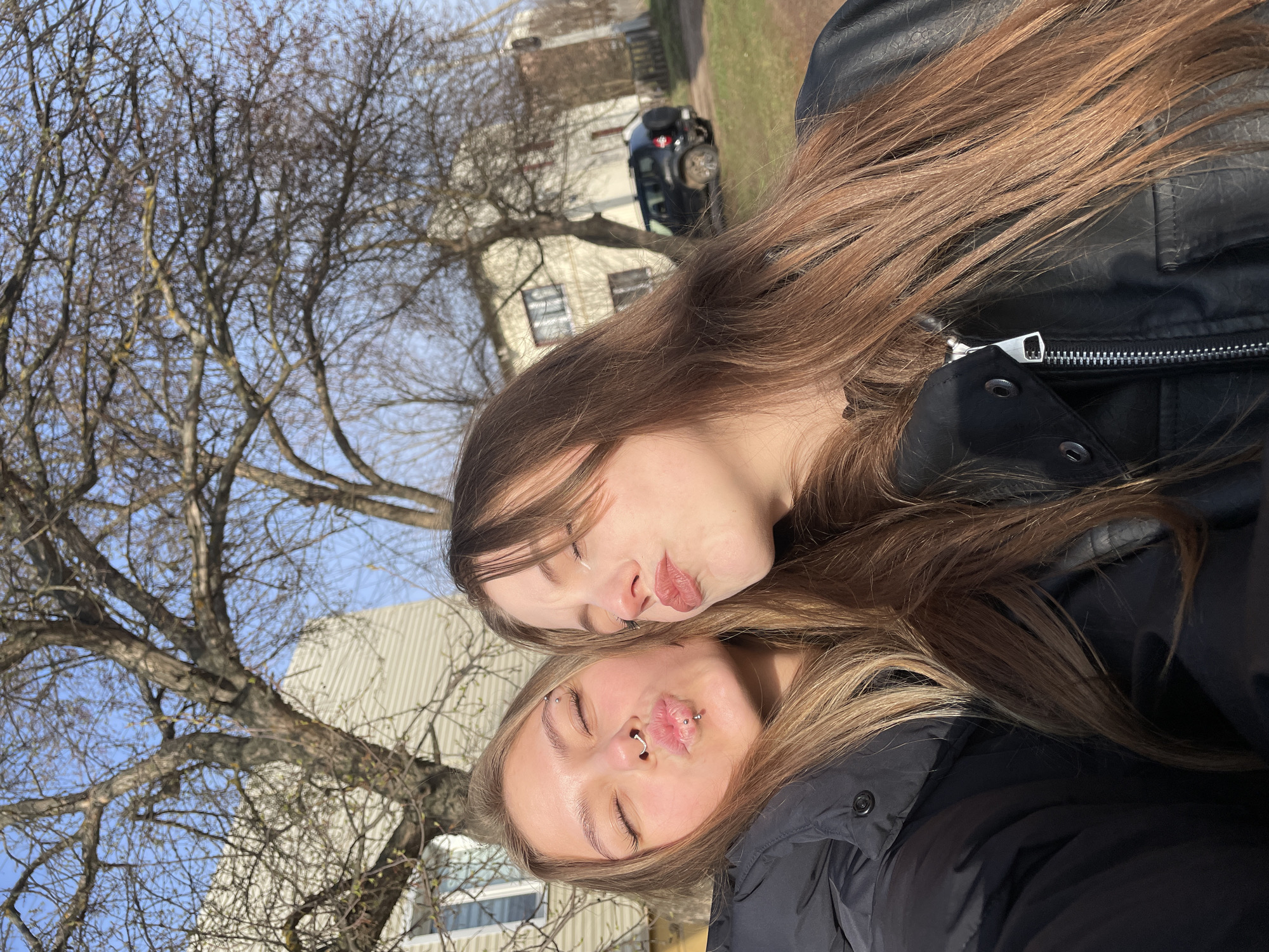 This rotated outdoor photograph captures two young women posing playfully, making "kiss" faces with their eyes closed, suggesting a selfie or a casual portrait. The image is oriented 90 degrees counter-clockwise from a standard portrait.
The woman positioned higher in the frame has long, straight, medium-brown hair and is dressed in a black jacket, likely leather, with a visible silver zipper. Her lips are softly pursed. Below her, the second woman features long, wavy, lighter brown or blonde hair and wears a black puffer jacket. She has several visible piercings: a septum ring, a philtrum piercing (medusa), and small studs or rings in both eyebrows. Her lips are also pursed in a matching pose.
The background reveals a sunny day with a clear blue sky. Dominating the middle ground are several large, deciduous trees with bare branches, suggesting the season is late autumn, winter, or early spring. In the distance, light-colored buildings, possibly houses or commercial structures, are visible. To the right, a dark-colored vehicle, appearing to be an SUV or jeep with a spare tire mounted on its rear, is parked on a patch of grass or dirt. The bright sunlight creates clear shadows, indicating it is likely mid-day. The scene depicts a casual moment in a semi-rural or suburban setting, identified as Фоки, Russia. No visible text is present in the image.