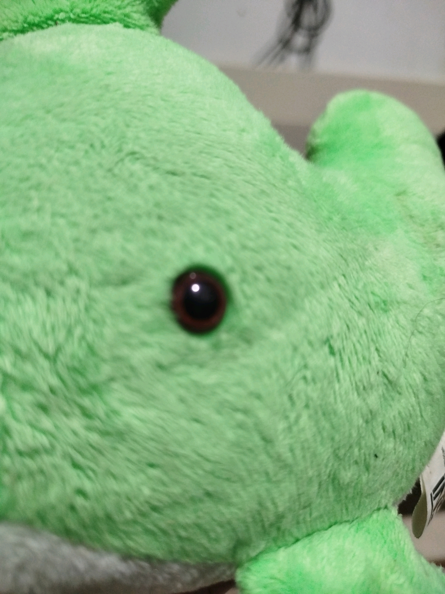 This is a close-up, shallow depth-of-field photograph of a plush toy. The toy is a vibrant green and appears to be made of a soft, fuzzy material. A single dark eye with a reflection is visible on the right side of the image, positioned on the plush toy's head. The background is out of focus, showing a blurred neutral-toned surface and what appears to be a faint sketch or drawing on a wall behind the toy. There are no people, actions, or interactions depicted. The location in Mojoagung, Indonesia, is not visually represented in the image.