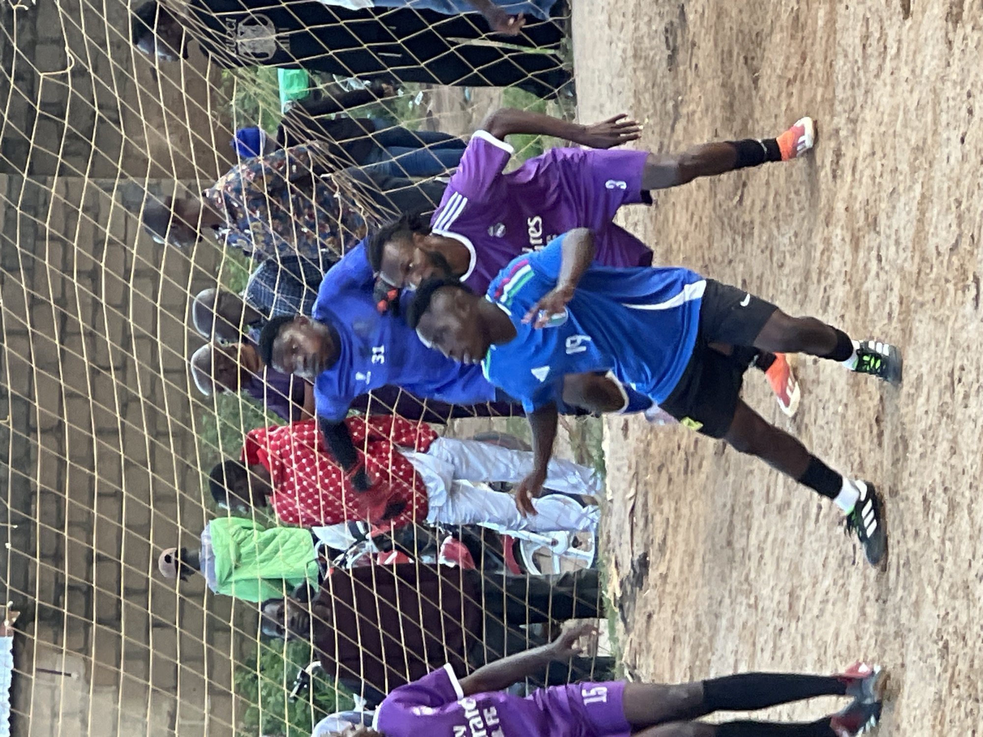 The image depicts a group of young men playing soccer on a dirt field, likely in Jos, Nigeria, evidenced by the presence of a brick wall and a chain-link fence, common in urban settings. In the foreground, a player in a blue jersey with white stripes and the number "19" on the front is in motion, his body angled as if about to kick or receive a ball. He is wearing black shorts and black soccer cleats with white stripes and green accents. To his right, a player in a purple jersey with white stripes and the number "3" on the shorts is also facing the action, his arms extended. Further left, another player in a purple jersey with the number "15" on the shorts is partially visible, wearing black socks and cleats. Behind the players and the net, several spectators are observing the game, including a person in a green jacket and a cap, another in a red and white polka dot shirt, and others in casual attire. The environment is dusty, with scattered debris on the ground. The overall scene suggests a lively, informal sporting event.