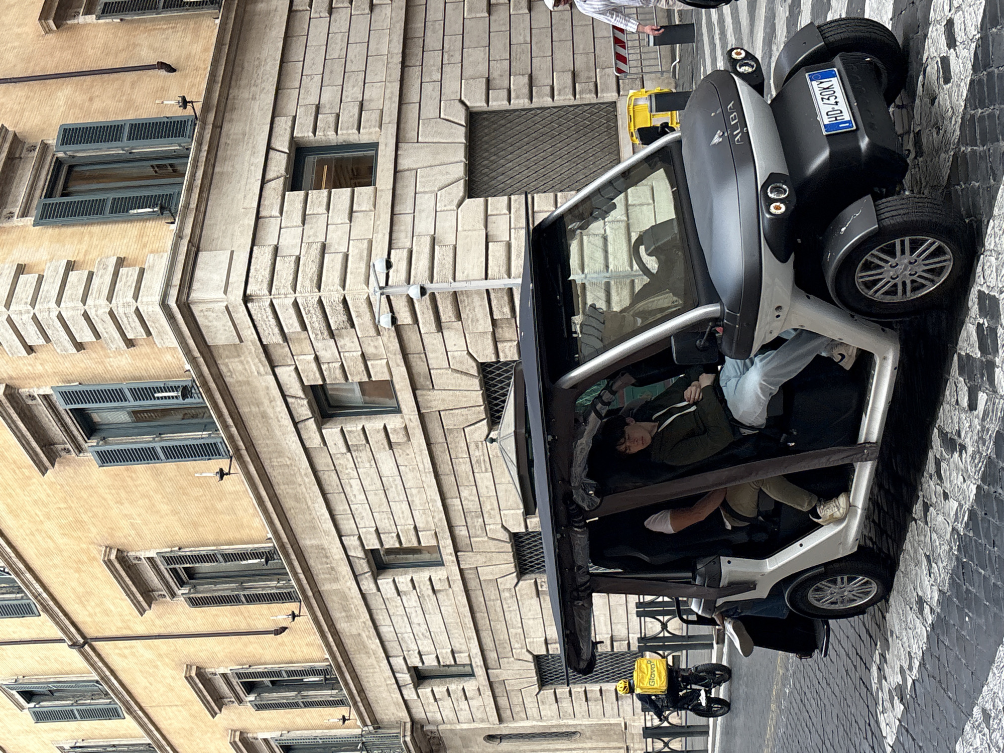 This image captures a vibrant street scene in Rome, Italy, dominated by a compact, two-seater electric vehicle. The car, which has "ALBA" emblazoned on its front and an Italian license plate "HD-430KY," is parked or temporarily stopped on a cobblestone street.
Inside the car, a passenger is seen reclining, possibly asleep or resting, wearing a dark top and light-colored trousers. Their body language suggests relaxation. The driver is partially visible, with only an arm and leg discernible. Near the front of the car, a person in a striped shirt and hat is standing, holding an object, possibly interacting with the vehicle or its occupants. Behind the car, a yellow "Glovo" delivery bag is visible on what appears to be a scooter or bicycle, suggesting a delivery person nearby.
The setting is characterized by historic European architecture. On the left, a building with a warm, light tan facade features multiple windows with traditional blue wooden shutters, some open, some closed. To the right, another building boasts a grey, textured stone facade with distinctive geometric patterns and smaller windows. The lighting indicates it's daytime, likely overcast, as there are no harsh shadows. The cobblestone street adds to the authentic Roman atmosphere.