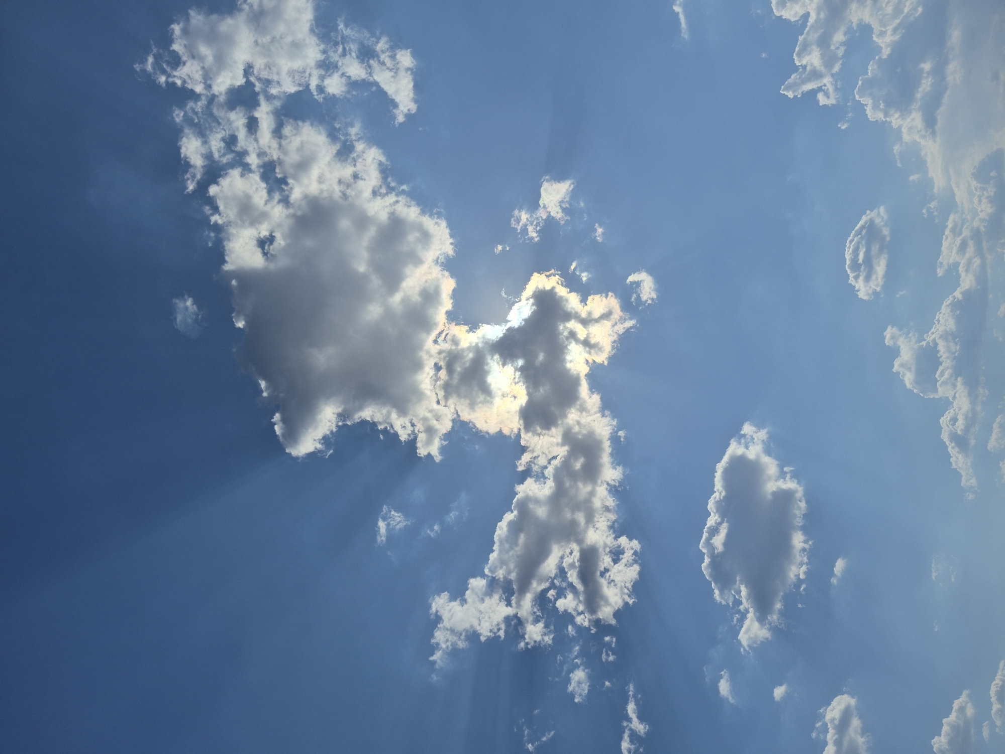 The image shows a blue sky with various white clouds. Sun rays are visible, emanating from behind a cluster of clouds in the center of the image. There are no people or objects other than clouds. The location is not specified.