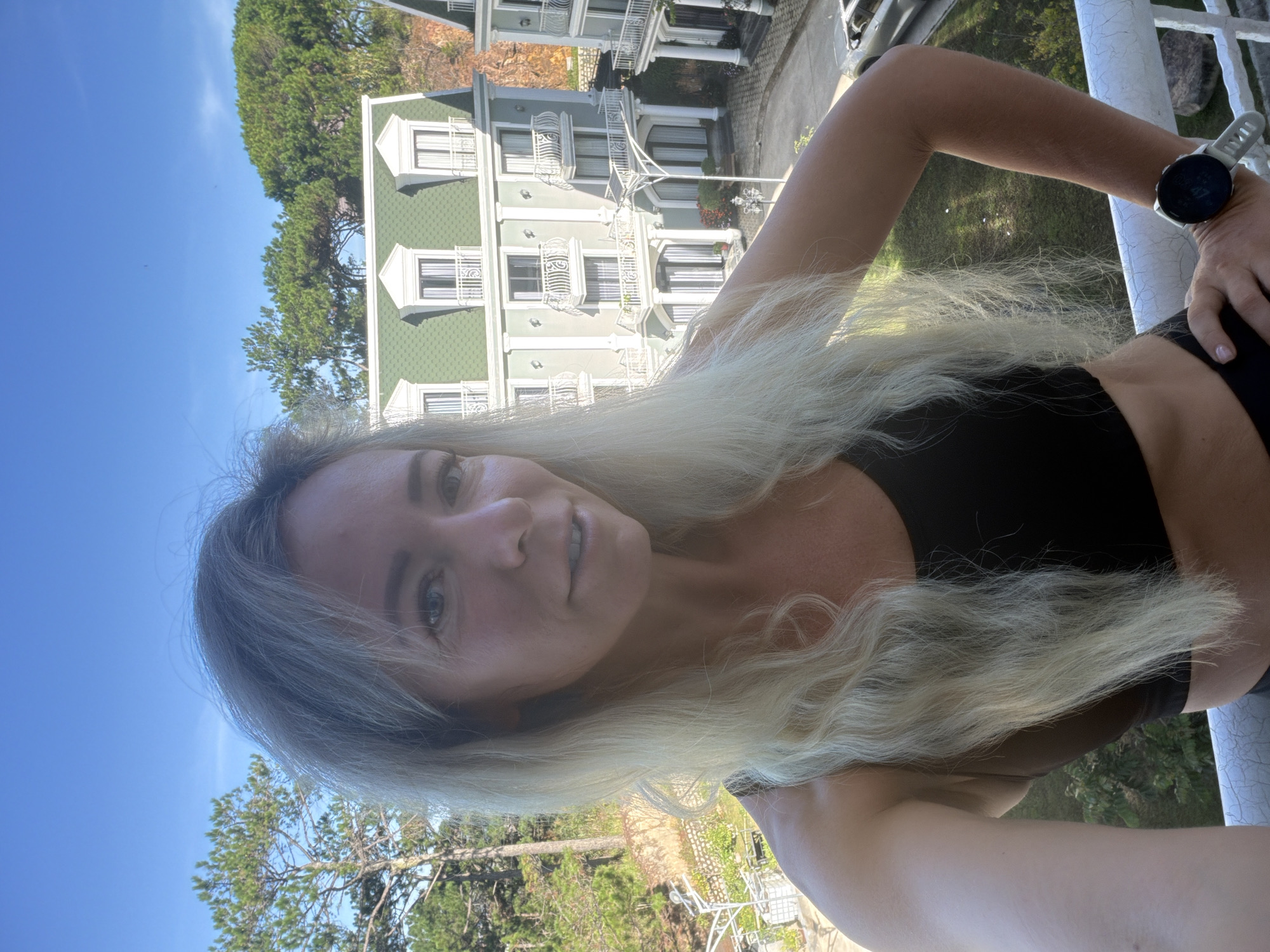 A blonde woman is standing on a balcony in front of a hotel in Xuan Truong, Da Lat, Vietnam. The hotel has a light green facade with white trim and balconies. The sky is blue and clear, with some trees visible in the background. The woman is wearing a black sports bra and black shorts. She has a fitness watch on her left wrist. She is looking at the camera and smiling. The overall impression is that of a tourist taking a selfie on vacation.