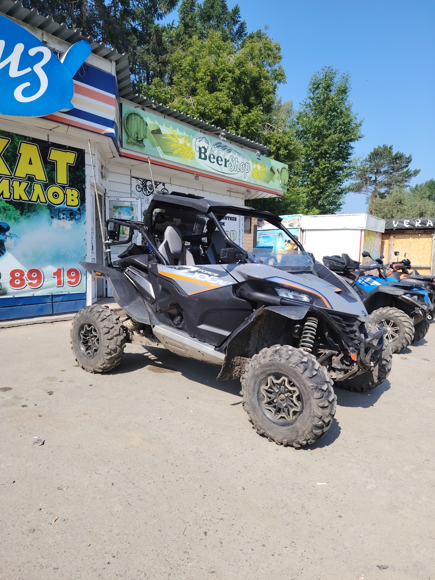 A side view of two all-terrain vehicles (ATVs) is captured in this outdoor photograph. The ATV in the foreground is a grey and black UTV with orange accents, parked on a paved surface. The ATV in the background is a blue ATV. Both vehicles are parked in front of a building with a corrugated metal roof. A sign above the building displays "BeerShop" with an image of a beer barrel and wheat stalks. Another sign to the left of the building shows "КАТ МИКЛОВ" in large yellow letters. The scene is set under a clear blue sky with green trees visible in the background.