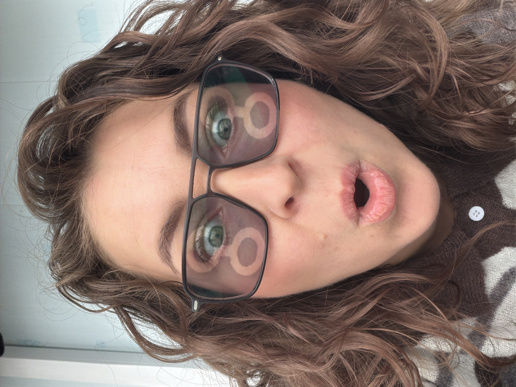 A close-up, rotated headshot captures a Caucasian female wearing dark-rimmed glasses. Her lips are slightly parted, giving the impression of blowing a kiss. Her eyes, visible through the glasses, are a light green color and reflect circular light sources. She has curly brown hair. She wears a brown and white patterned sweater with a white button. The background appears to be a white wall or surface. The location of the image is unknown.