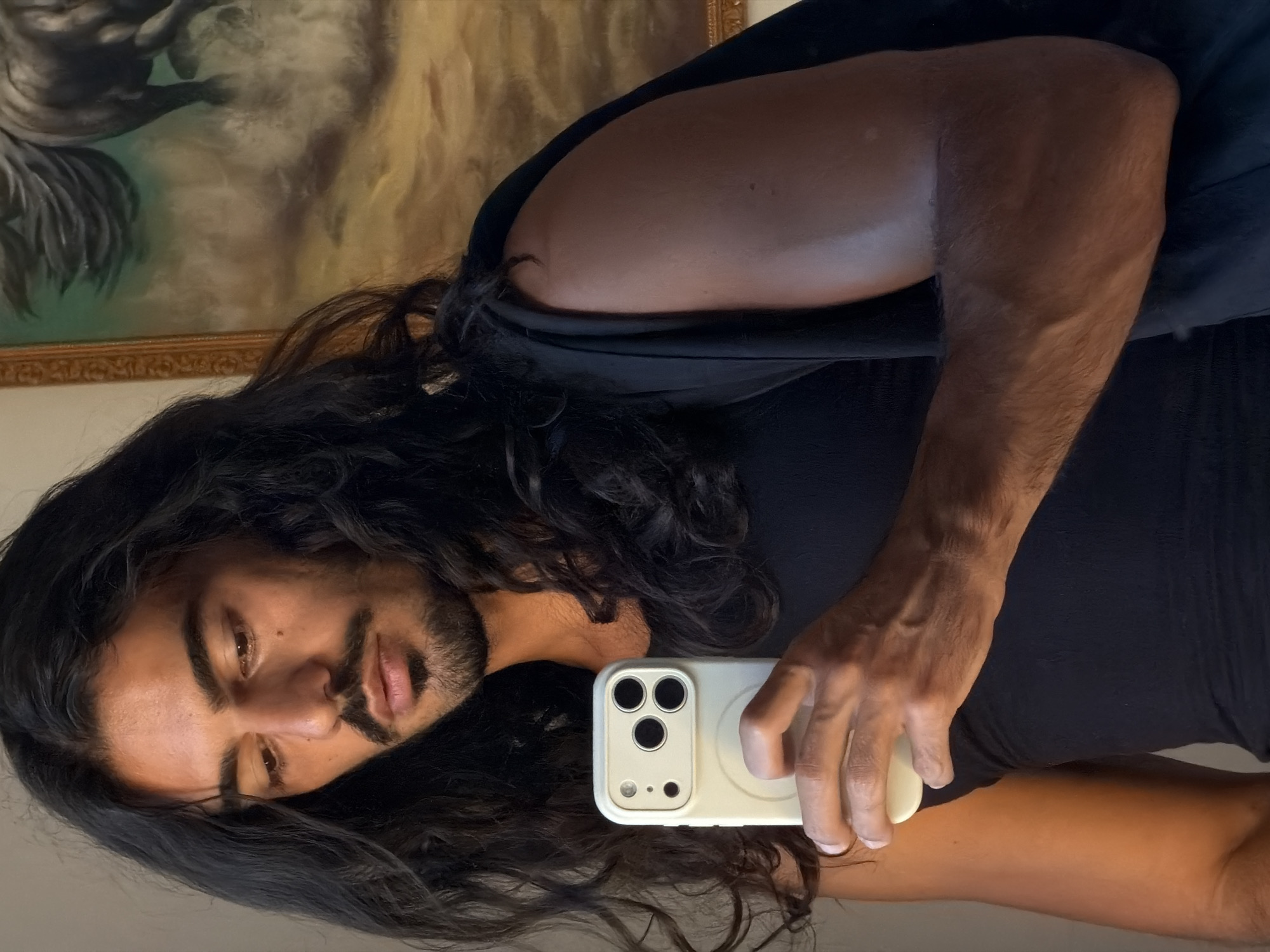 A man with long, dark, wavy hair is taking a selfie. He is wearing a black tank top. His left arm is raised holding a white smartphone with a triple-lens camera. A painting with a gold frame is visible in the background. The image was captured in North Kuta, Indonesia.