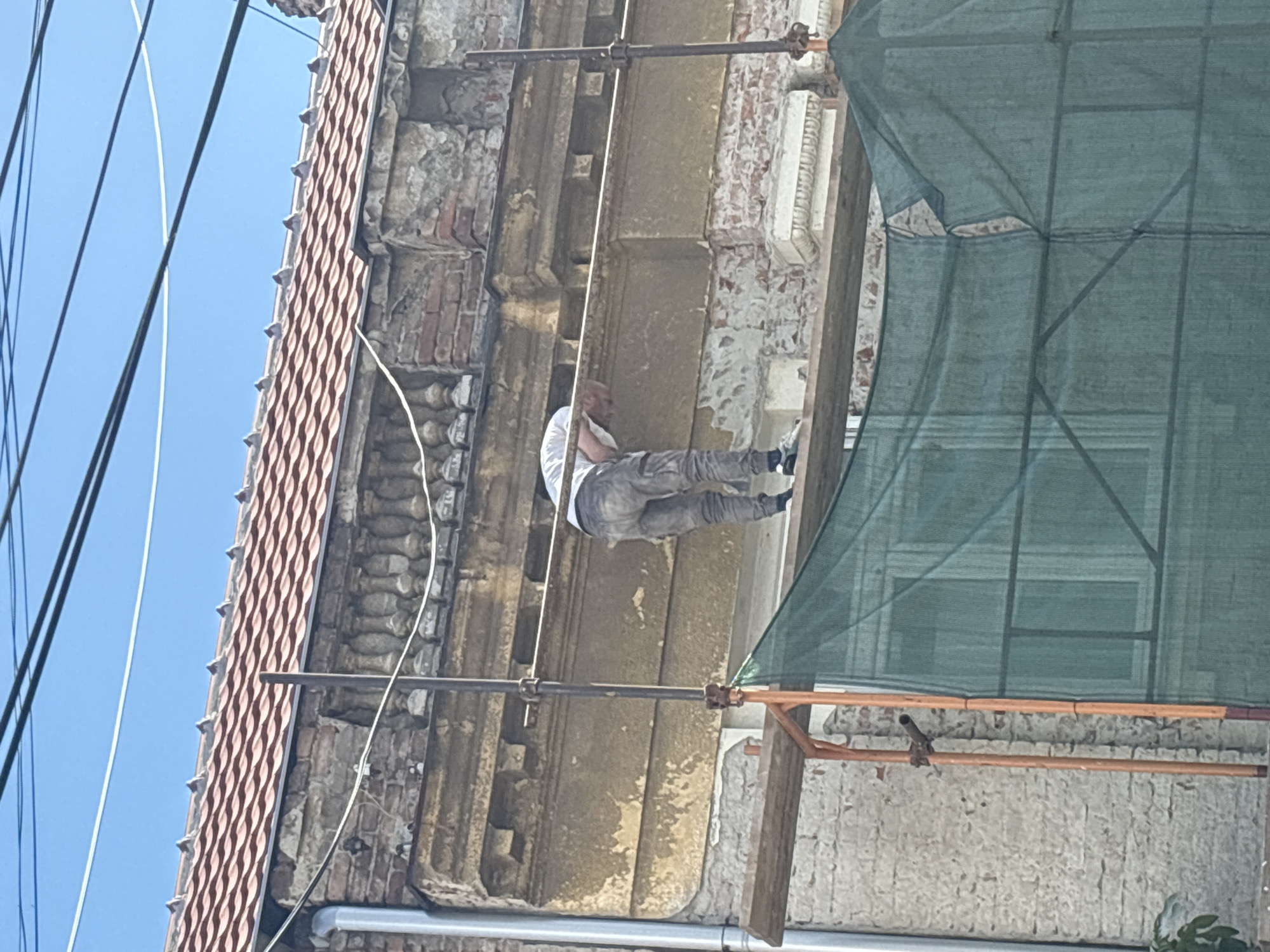 An individual, wearing a light-colored t-shirt and gray work pants, is performing maintenance on the facade of a building. They are positioned on a wooden plank supported by scaffolding. The building exhibits weathered brickwork and decorative stone elements, suggesting it is undergoing renovation. The scaffolding is covered with a green mesh tarp. The background consists of a clear blue sky and overhead electrical wires. This scene is likely located in the Vracar Urban Municipality in Serbia, a region known for its historic architecture and ongoing urban development.