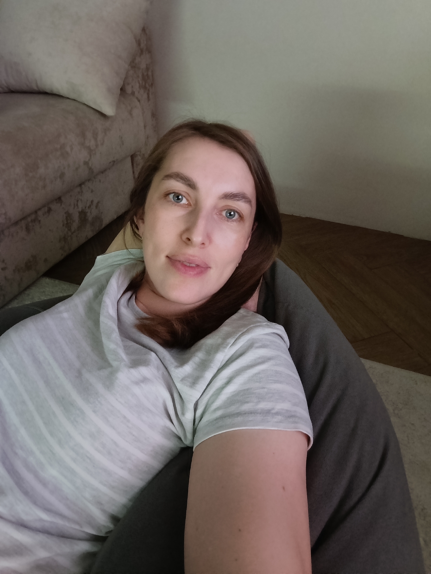 A woman with fair skin and blue eyes is reclined on a dark grey beanbag chair. She is wearing a grey and white striped t-shirt. Her brown hair is parted to the side. A tan colored couch is partially visible in the upper left corner. Hardwood flooring is visible in the upper right corner. The woman is looking directly at the camera. The location is unknown, but is presumed to be in the city of Chaikovsky, Russia.