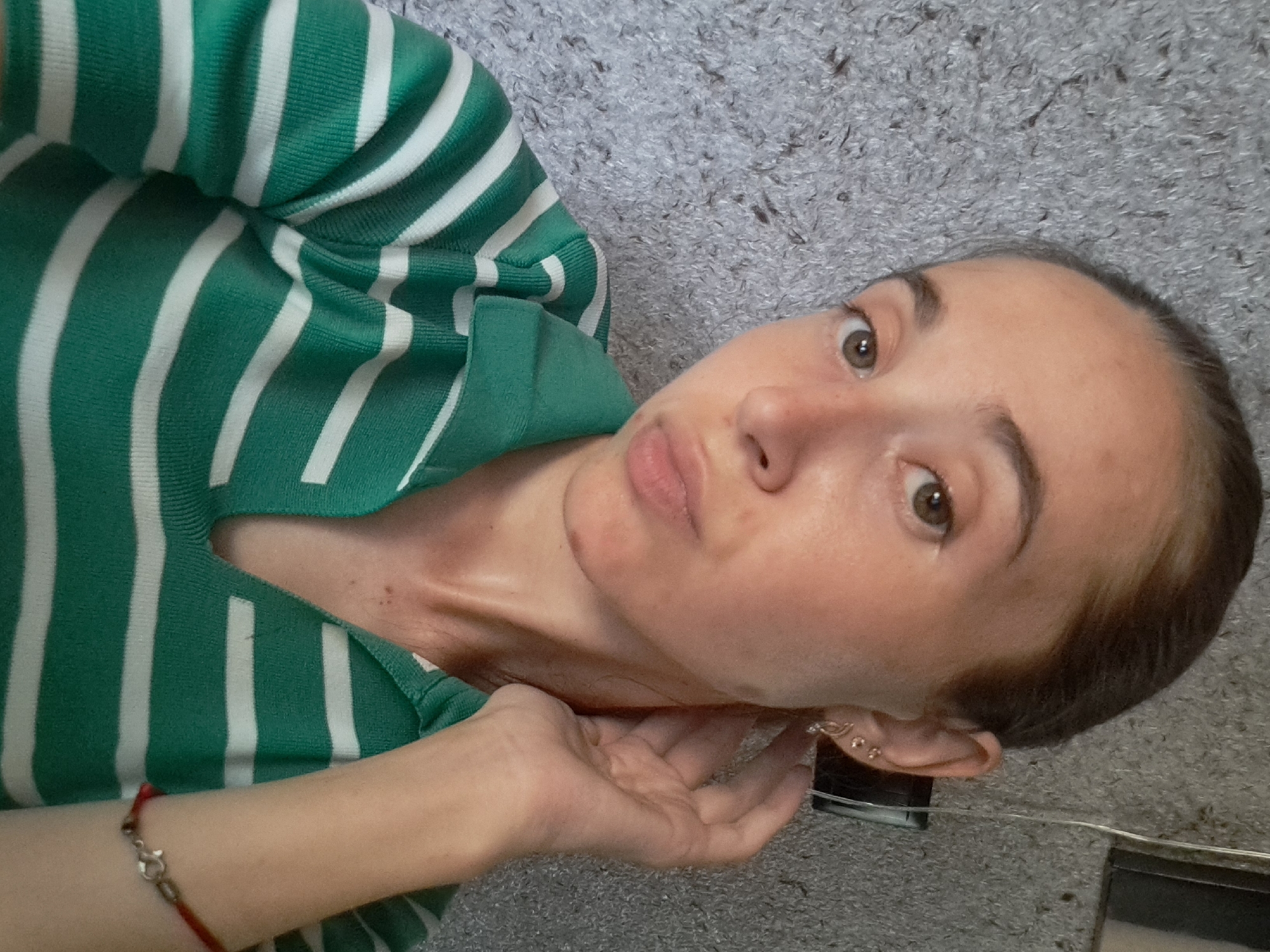 In Vypasne, Ukraine, a young woman is captured in a self-portrait. She has fair skin, brown eyes, and her dark hair is pulled back. She is wearing a green and white striped polo shirt and a delicate earring in her left ear. Her right hand is raised to her ear, and a thin red bracelet adorns her wrist. The background is a textured gray wall.