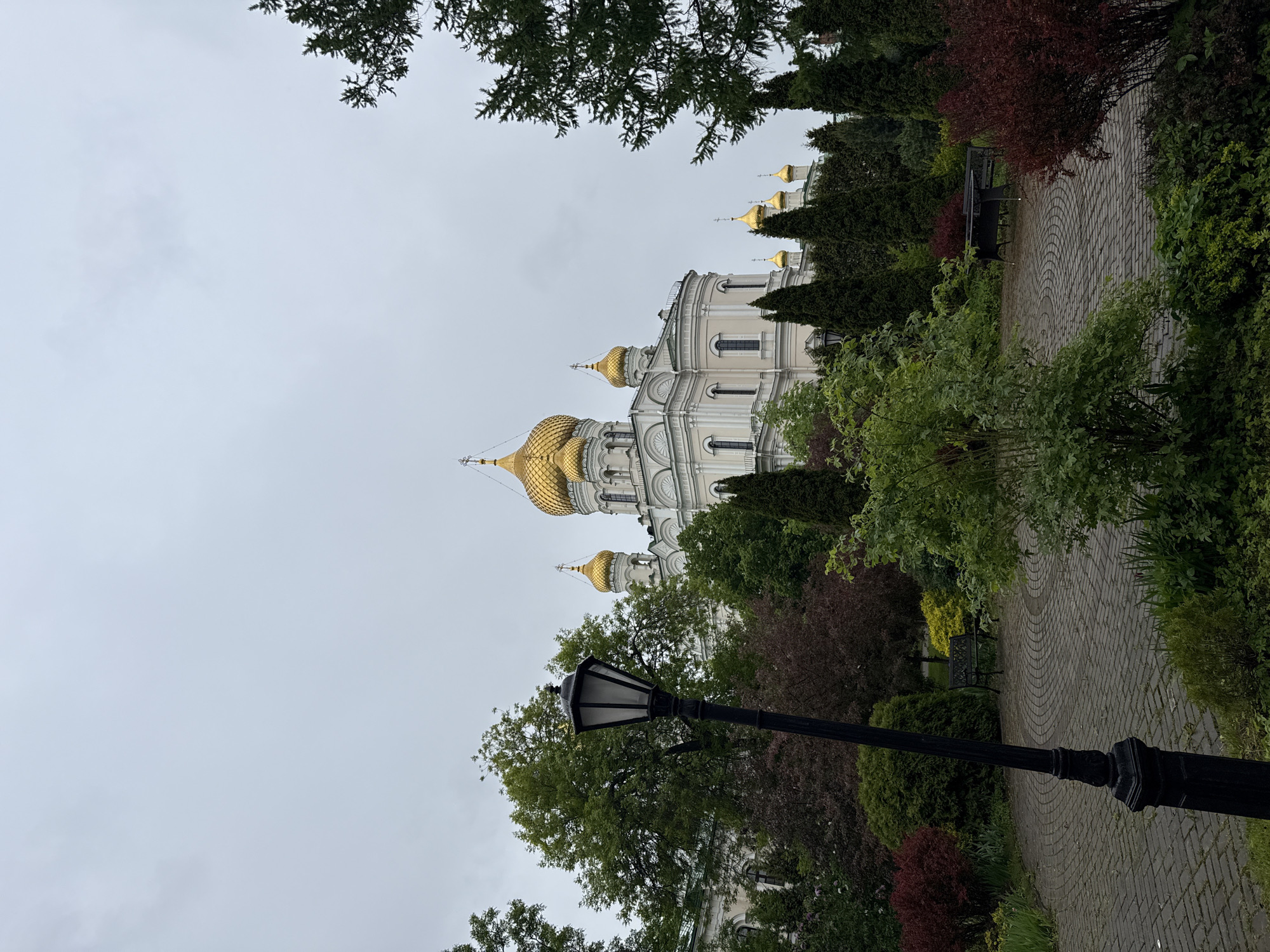 The image depicts the St. John Convent in Saint Petersburg, Russia. The architecture of the convent building is visible, featuring golden domes and a white facade. The convent is surrounded by greenery, including trees, bushes, and a paved walkway. A black lamppost is in the foreground, partially obscuring the view. The sky is overcast. A bench can be seen on the walkway.