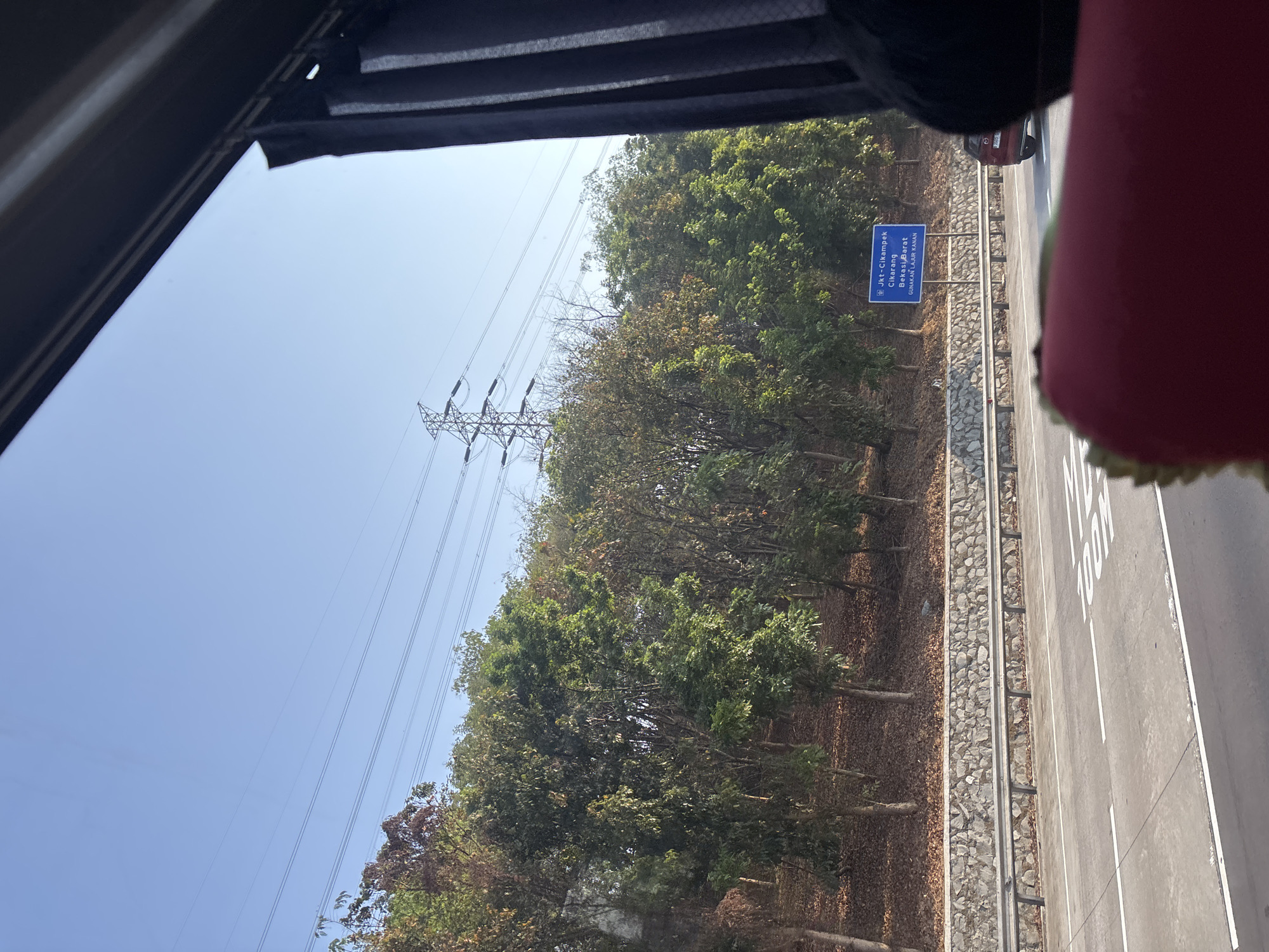 The image shows a view from inside a vehicle on a highway, likely in Karawang, Indonesia, given the signage. The highway has multiple lanes with white markings. A blue signpost on the side of the road indicates destinations: "Jkt-Cikampek," "Cikarang," and "Bekasi Barat," with instructions to "Gunakan Lajur Kanan" (Use the Right Lane). Trees line the embankment behind a stone retaining wall. In the distance, a tall electricity pylon with high-voltage power lines stretches across the clear blue sky. A red car is visible in the right lane. The foreground is partially obscured by a red cushioned object, possibly a seat or accessory within the vehicle. The lighting suggests it is daytime.