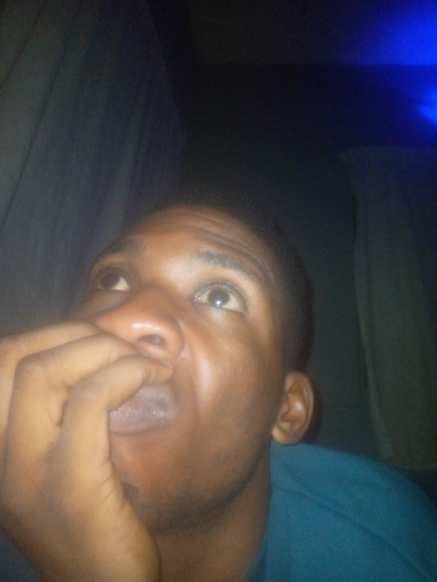 A close-up shot of a man with dark skin, a short haircut, and a teal t-shirt. He is looking up and to the left, with his hand partially covering his mouth. The background includes what appears to be a dark curtain or wall on the left, and another, slightly lighter, drape or wall on the right. There is a patch of blue light visible in the upper-right. The location is Unknown, Unknown.