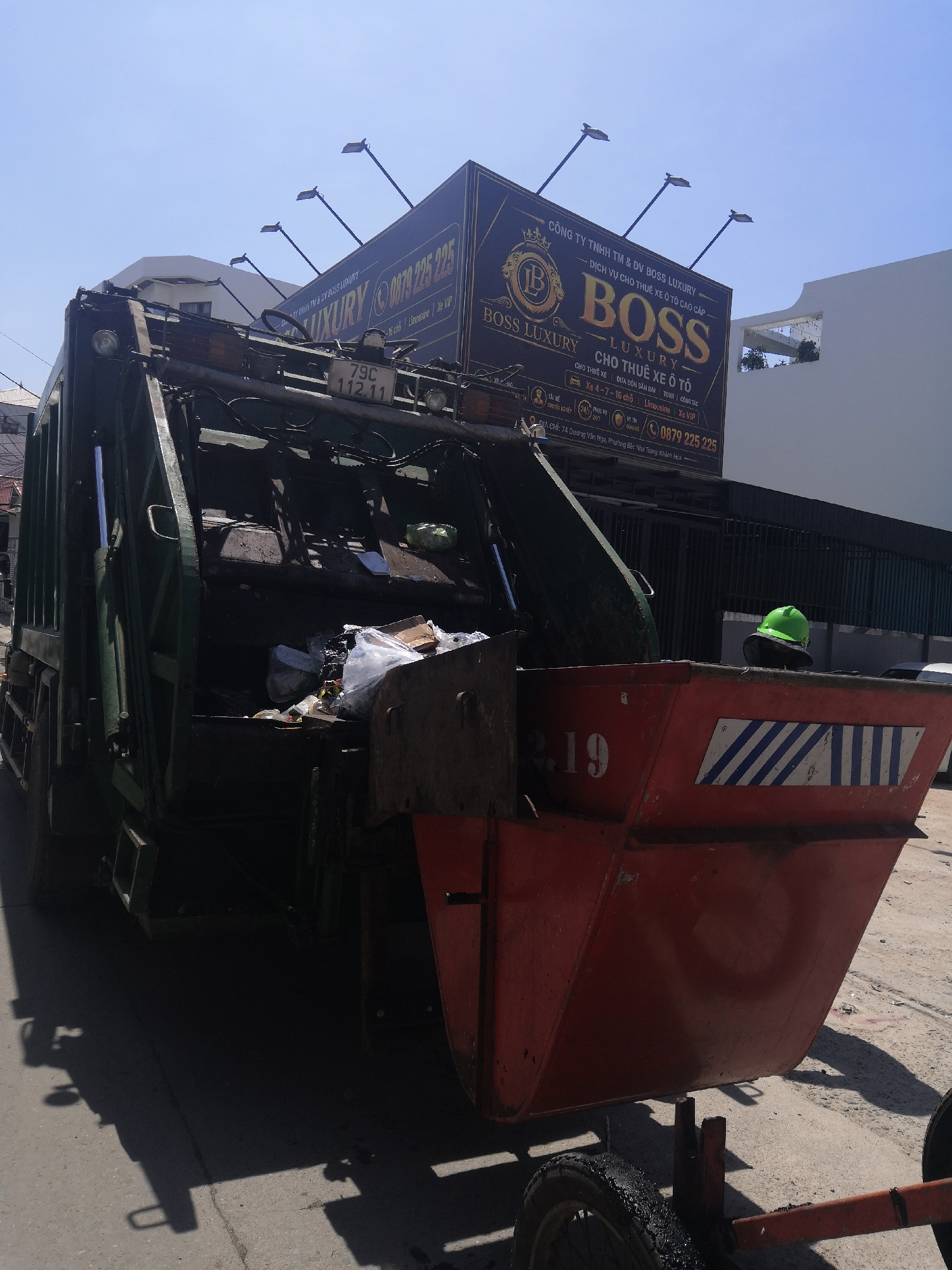 A dark green garbage truck is parked on a paved street under a clear blue sky. The truck's rear hopper is open, revealing various types of refuse, including plastic bags and cardboard. Attached to the rear of the truck is a reddish-orange wheeled dumpster, also filled with trash. A bright green safety helmet rests on top of the refuse within the dumpster. The dumpster features white and blue diagonal stripes as a reflective marker and the number "2.19" inscribed on its side. The truck's license plate, "79C 112.11", is visible above the open hopper. Dominating the upper background is a large black and gold billboard advertising "CÔNG TY TNHH TM & DV BOSS LUXURY" (BOSS LUXURY TRADING & SERVICES CO., LTD) for "DỊCH VỤ CHO THUÊ XE Ô TÔ CAO CẤP" (High-End Car Rental Service). The advertisement lists services such as car rental, airport transfer, tours, business trips, and offers 4-7-16 seater cars, limousines, and VIP cars, with a contact number "0879 225 225". The billboard specifies the address as "74 Dương Vân Nga, Phường Bắc Nha Trang, Khánh Hòa", confirming the location in Bac Nha Trang Ward, Vietnam. Three streetlights are visible on poles extending upwards from behind the billboard. Modern white buildings are present in the background to both sides of the billboard. Strong sunlight from the right casts defined shadows on the pavement.