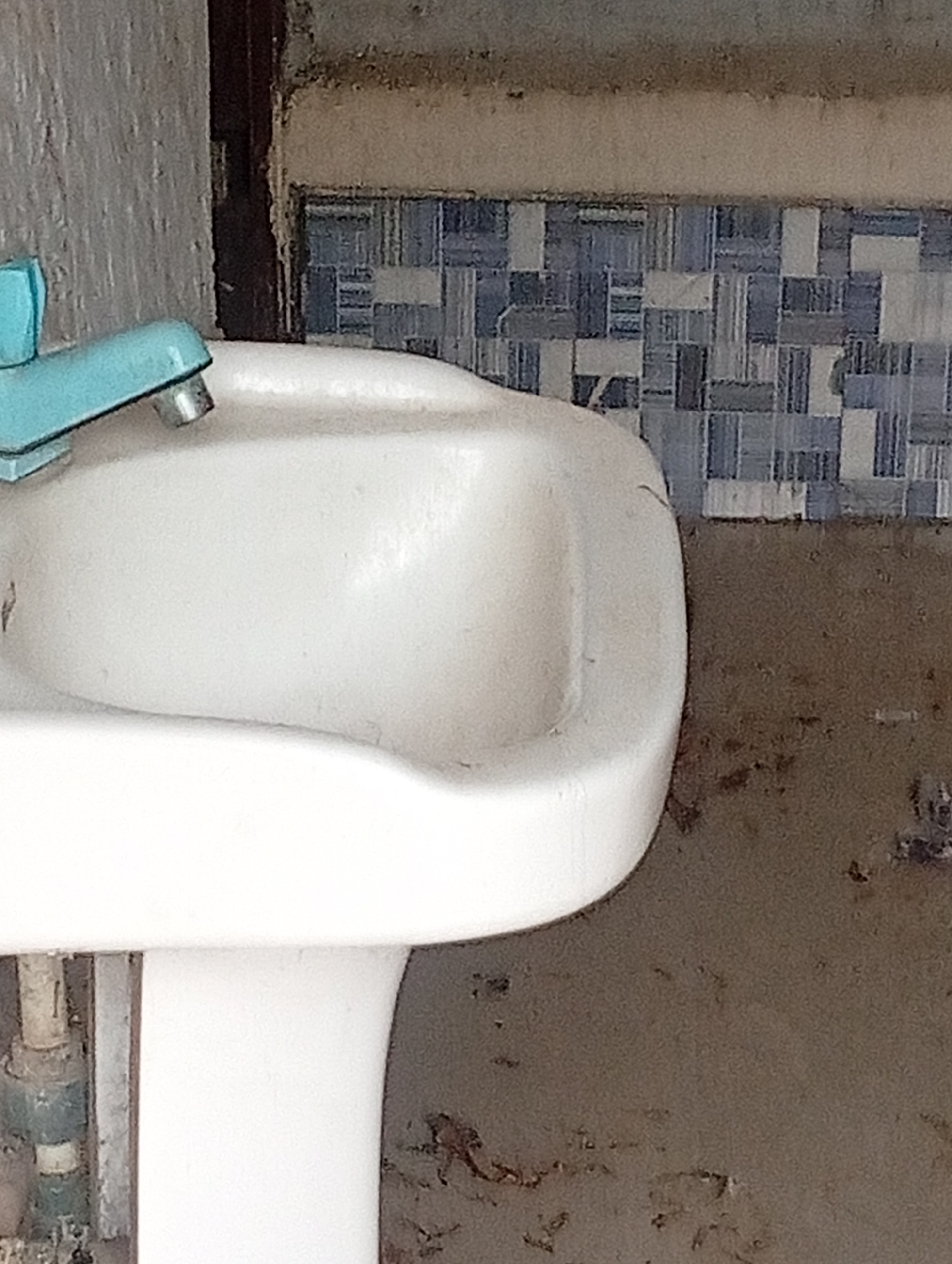 A close-up, eye-level, medium shot shows a portion of a white porcelain sink with a teal-colored faucet. The sink is positioned in what appears to be a bathroom setting. Behind the sink, a tiled wall features a pattern of blue and white rectangular tiles. The wall above the tiles is a muted beige color with some discoloration and grime. To the left of the sink, a textured grey wall is visible. The lower portion of the image shows the base of the sink pedestal with some plumbing pipes. The environment appears somewhat aged and potentially in need of cleaning, with visible marks and stains on the walls. The location is identified as Jalingo, Nigeria.