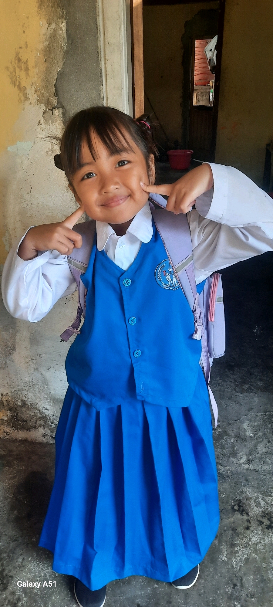 A young girl, likely of Southeast Asian descent, stands in a doorway, facing the camera and smiling. She is wearing a school uniform consisting of a white collared shirt, a blue vest, and a blue pleated skirt. A light purple backpack with pink accents is visible over her shoulders. She is playfully touching her cheeks with her index fingers. The background shows a rough, weathered wall to her left and an open doorway to her right, revealing a dimly lit interior space with a red bucket and some wooden structures. The floor appears to be concrete. The image has a timestamp at the bottom left corner indicating "Galaxy A51". The location is identified as Ploso, Indonesia.