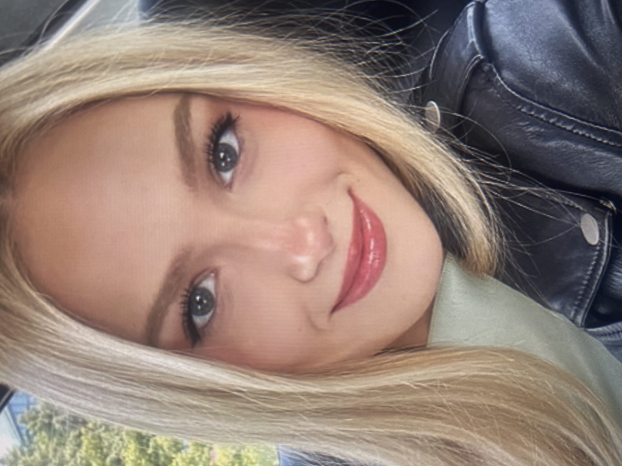 A close-up, medium shot captures a young Caucasian woman from the shoulders up, seated in a vehicle. She has blonde hair that falls past her shoulders and is styled to frame her face. Her eyes are a striking blue-gray, accentuated with dark eyeliner and mascara. Her eyebrows are well-defined and arched, and she has a light dusting of blush on her cheeks. Her lips are full and painted with a glossy, rosy-pink lipstick. She is wearing a light green top beneath a black leather jacket, which features prominent stitching and a collar. The background is blurred, showing hints of green foliage and a structure that suggests an outdoor setting within an urban environment. The image has a slightly pixelated quality, consistent with a digital photograph.