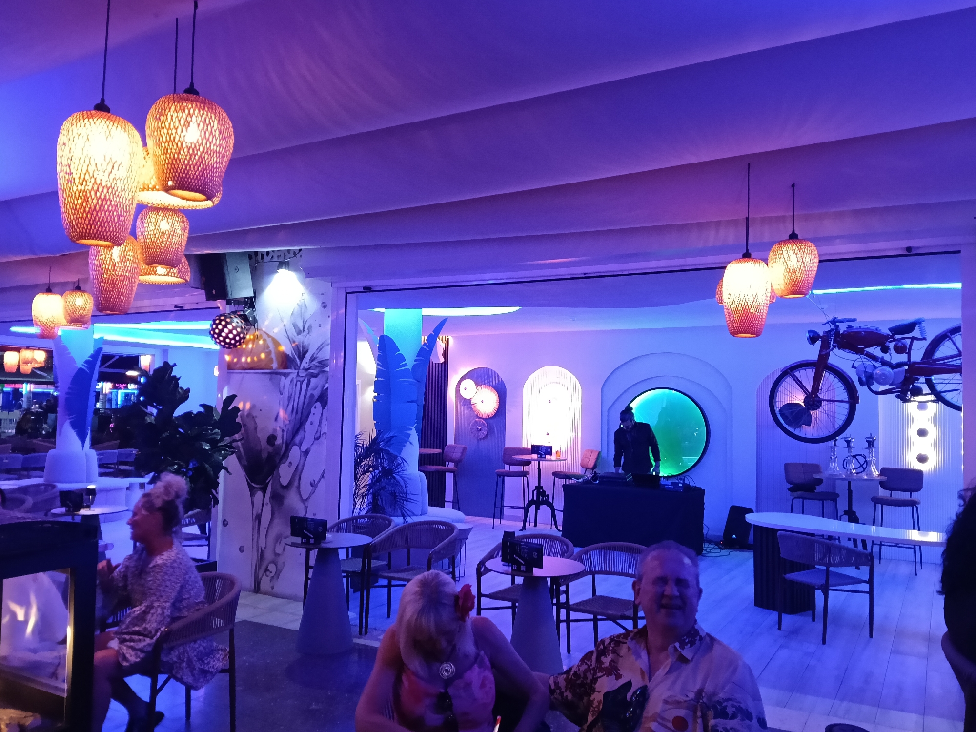 A nighttime scene in a bar setting in Playa Del Inglés, Spain. There are five patrons and one DJ. The DJ stands behind his equipment in the center of the image. The interior of the bar features several seating areas with small tables and chairs. Decorative lighting fixtures hang from the ceiling. A decorative motorcycle hangs on the wall. The interior design includes arched doorways and artistic wall features. The overall color tone is blue and purple.