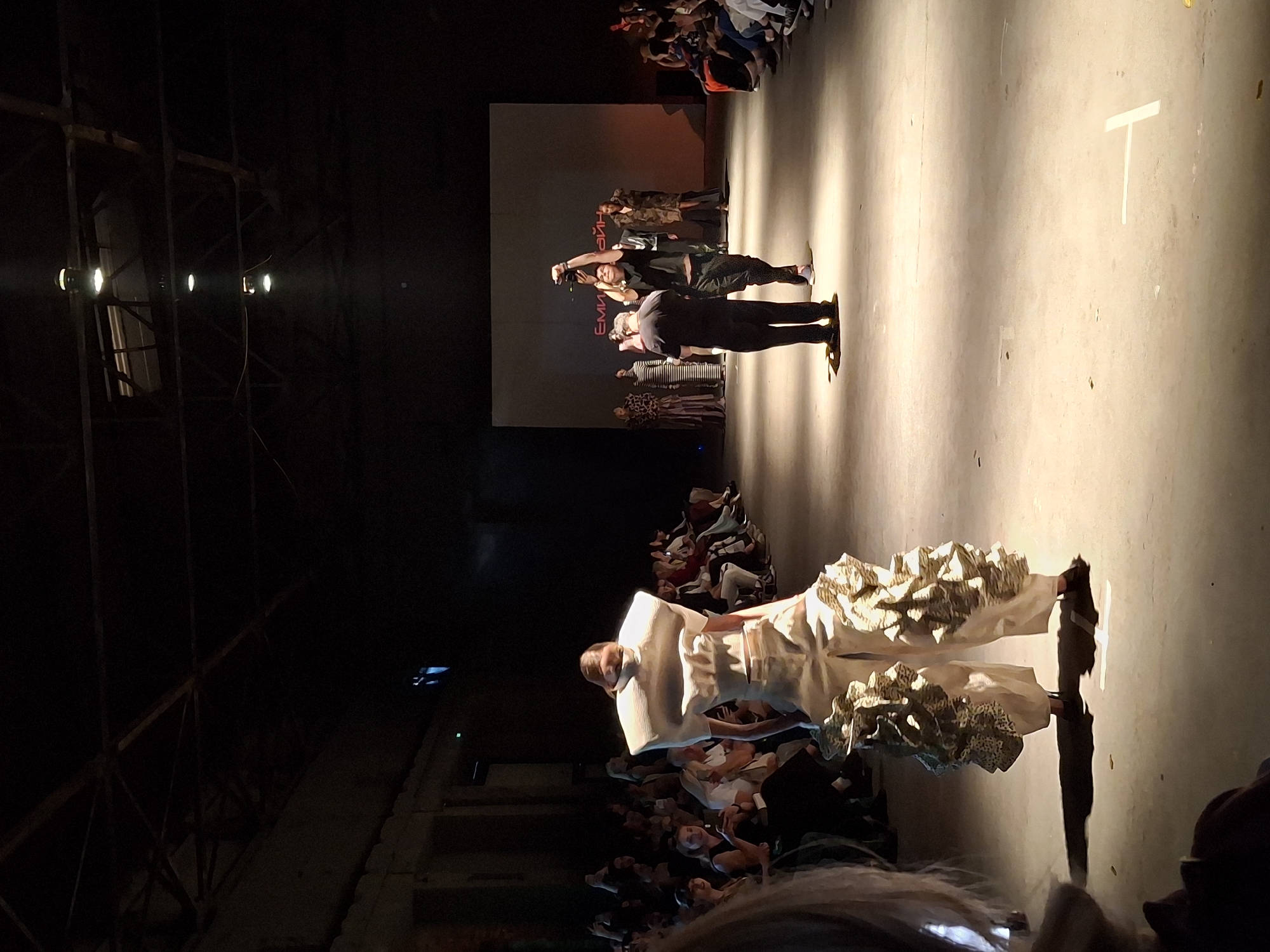The image shows a fashion show. There are people sitting in the audience. A person in an elaborate white outfit is walking on the runway. At the back, there are people onstage and behind them is a screen with text.
The location is Unknown, Unknown.
