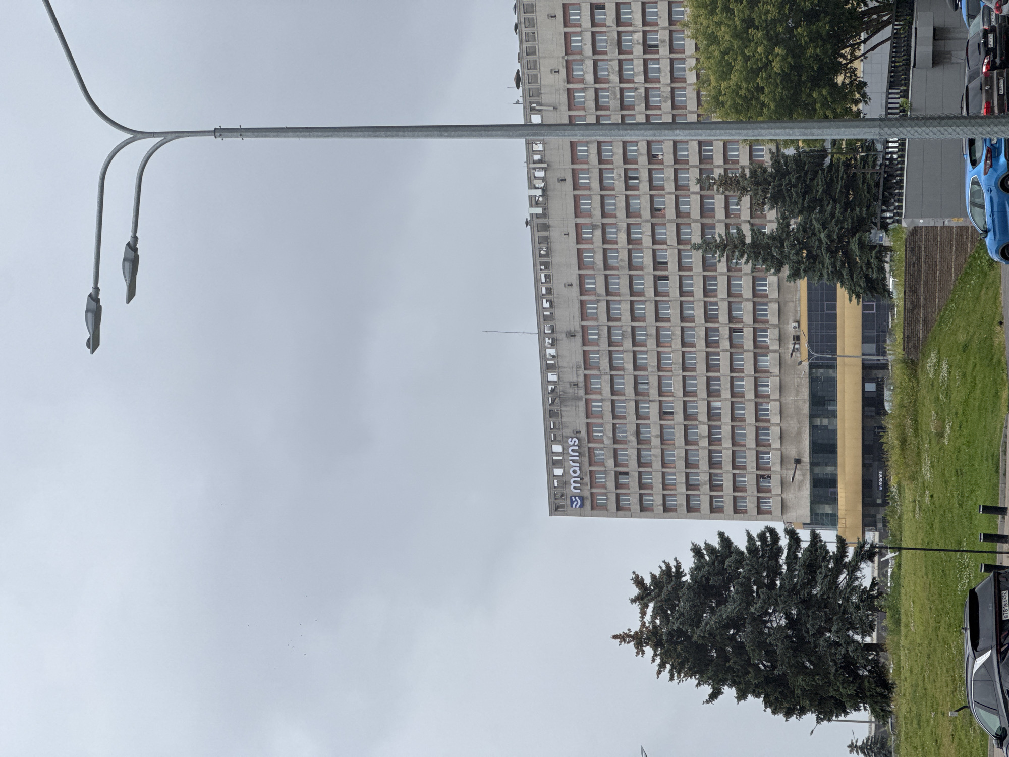 The image depicts an overcast sky over a city scene in Nizhny Novgorod, Russia. In the foreground, a grey metal streetlamp with two lights extends upwards and branches out, suggesting it's daytime, though the cloudy weather creates a subdued light.
Dominating the mid-ground are two imposing, multi-story buildings. The first, on the left, is a light-colored, Brutalist-style structure with rows of uniform windows. The second, to its right, shares a similar architectural style, also featuring numerous windows. A prominent sign on this building displays the word "marins" in blue lettering, accompanied by a blue logo. Between the buildings, a dark green coniferous tree stands tall.
To the right of the buildings, a strip of bright green grass borders a paved area where several cars are parked. A blue car is clearly visible, alongside other vehicles, one of which is dark and has a license plate ending in "BK 352". The overall setting appears to be an urban environment, possibly a business district or residential area, with the presence of streetlights, buildings, and parked cars. The weather is clearly cloudy, contributing to a muted and possibly cool atmosphere. There are no people visible in the frame, and no specific event is clearly taking place; it's a static street view.