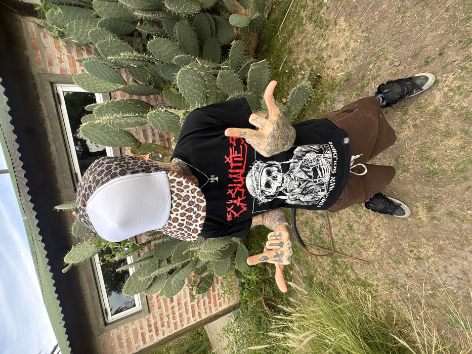 A lone individual is depicted in an outdoor, high-angle, full-body portrait. The subject stands on dry, patchy ground composed of dirt and sparse green grass, with some taller feathery grasses to the left. Their face is entirely obscured by a white-fronted, leopard-print trucker hat. They wear a black t-shirt with red text "THE CASUALTIES" above a black-and-white graphic of a skeleton in a military helmet with a crown of thorns, holding a rifle, accompanied by the text "UNKNOWN SOLDIER." Brown drawstring shorts and black and white athletic shoes are worn. The subject exhibits extensive tattoos on the neck, both arms, and hands, with "13" visible on the knuckles of the left hand and an eye design on the right hand. Large ear gauges are present in both earlobes. The left hand is extended outwards with the thumb and index finger raised, while the right hand is extended with the middle finger raised.
In the background, a single-story structure features red brick walls, a white-framed rectangular window, and a dark green corrugated metal roof. A dense cluster of large, green Opuntia cacti with numerous pads and spines grows directly behind the subject and against the brick wall. A bent, dark metal object lies on the ground near the subject's left foot. The sky overhead is overcast. The specific location within the city of Buenos Aires, Argentina, cannot be determined from the visual information provided.