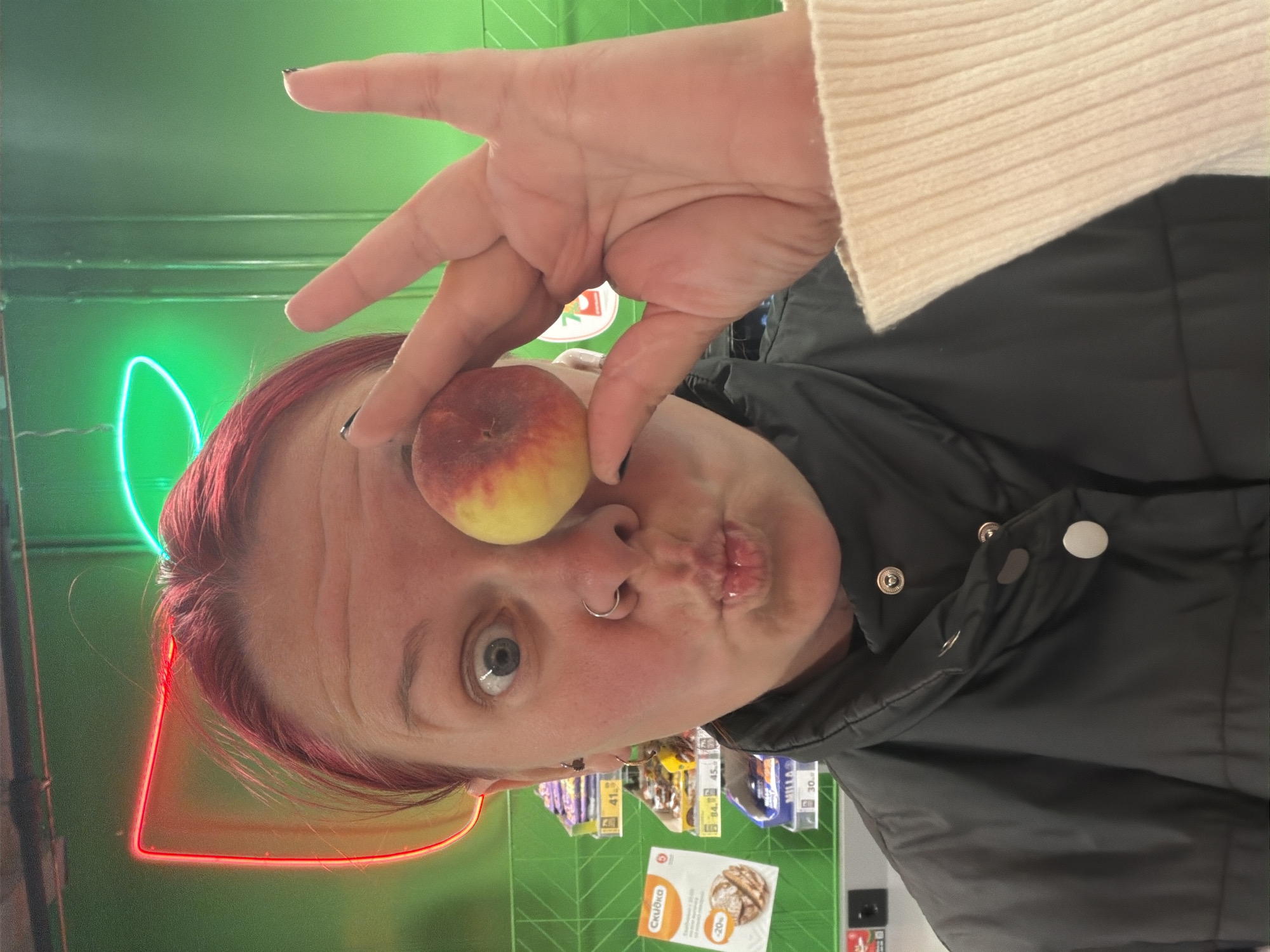 In a brightly lit store, possibly a supermarket in Chaikovsky, Russia, a person with vibrant red hair and a nose ring is playfully holding a peach over their right eye. Their left eye is wide open and looking towards the camera, while their lips are pursed in a kiss-like expression. They are wearing a black puffer jacket and a cream-colored ribbed sweater. Behind them, shelves are stocked with various products, some labeled with Cyrillic text and prices, and a neon sign glows in the background, casting a green and red light. The person's right hand, with painted black nails, is raised, holding the peach and making a peace sign with their index and middle fingers. The overall scene suggests a lighthearted moment captured within a retail environment.