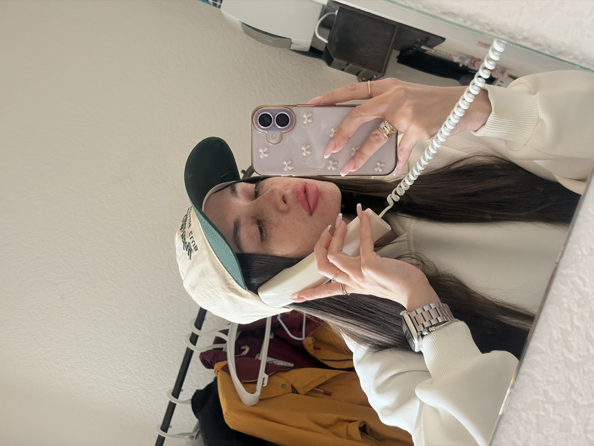A woman in a mirror selfie is holding a landline phone to her ear and a smartphone in her other hand. She is wearing a green and beige baseball cap and a white sweatshirt. Her hair is dark and straight. She has a silver watch on her left wrist. There is a clothes rack with various colored shirts visible in the reflection. The background is a white wall. The location in Saint Petersburg, Russia is unknown.