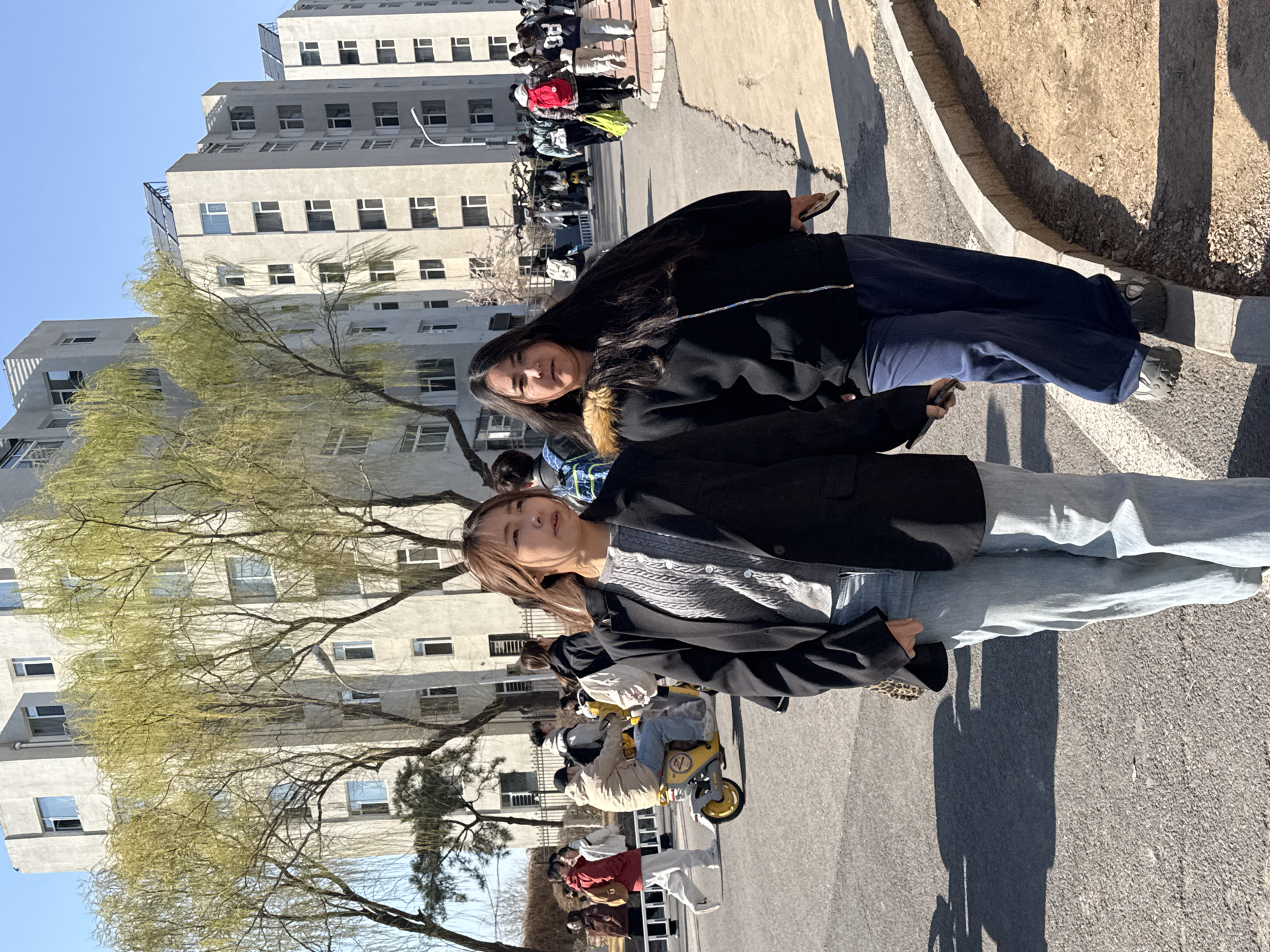 Two young women stand posing in Zhongtun, China, on a sunny day. The woman in the foreground has short brown hair and wears a black jacket, a gray knit sweater, and light blue jeans. The woman behind her has long black hair and wears a black jacket with fur lining and dark blue pants. Behind the women, there is a line of people, parked bikes, and a tall, gray, multi-story building. Trees with sparse green leaves are also visible. The location is a paved area, possibly a street or sidewalk.