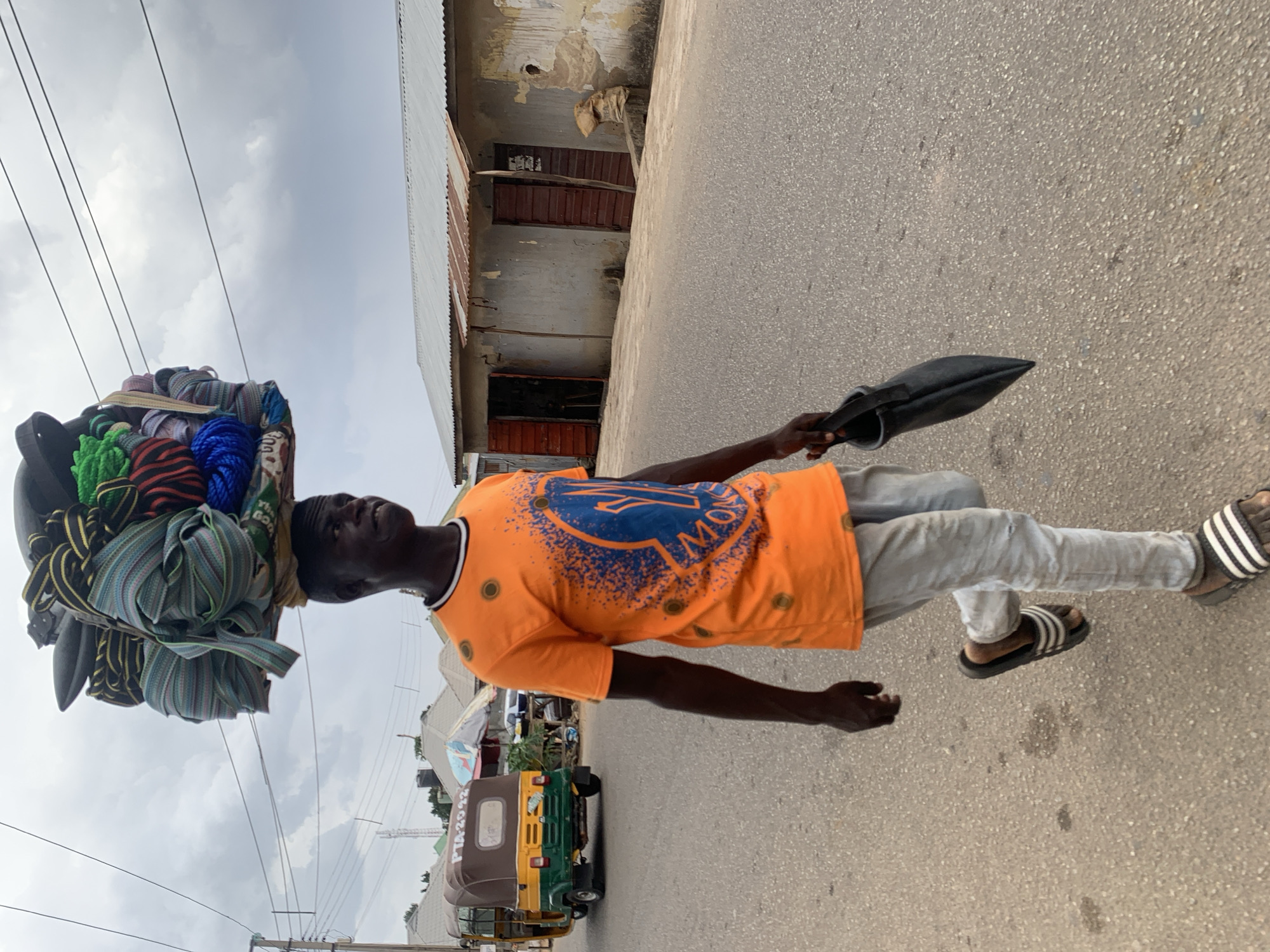 A man walks along a paved road in Jos, Nigeria. He carries a large bundle of goods, including ropes and striped fabrics, balanced on his head. In his hand, he holds a dark, cone-shaped bag. The man wears an orange t-shirt with a blue design, light-colored pants, and black and white striped sandals. Behind him, a green and yellow three-wheeled vehicle is parked. A building with closed brown shutters and a corrugated metal roof is visible to the right of the man. Power lines stretch across the sky above the scene.