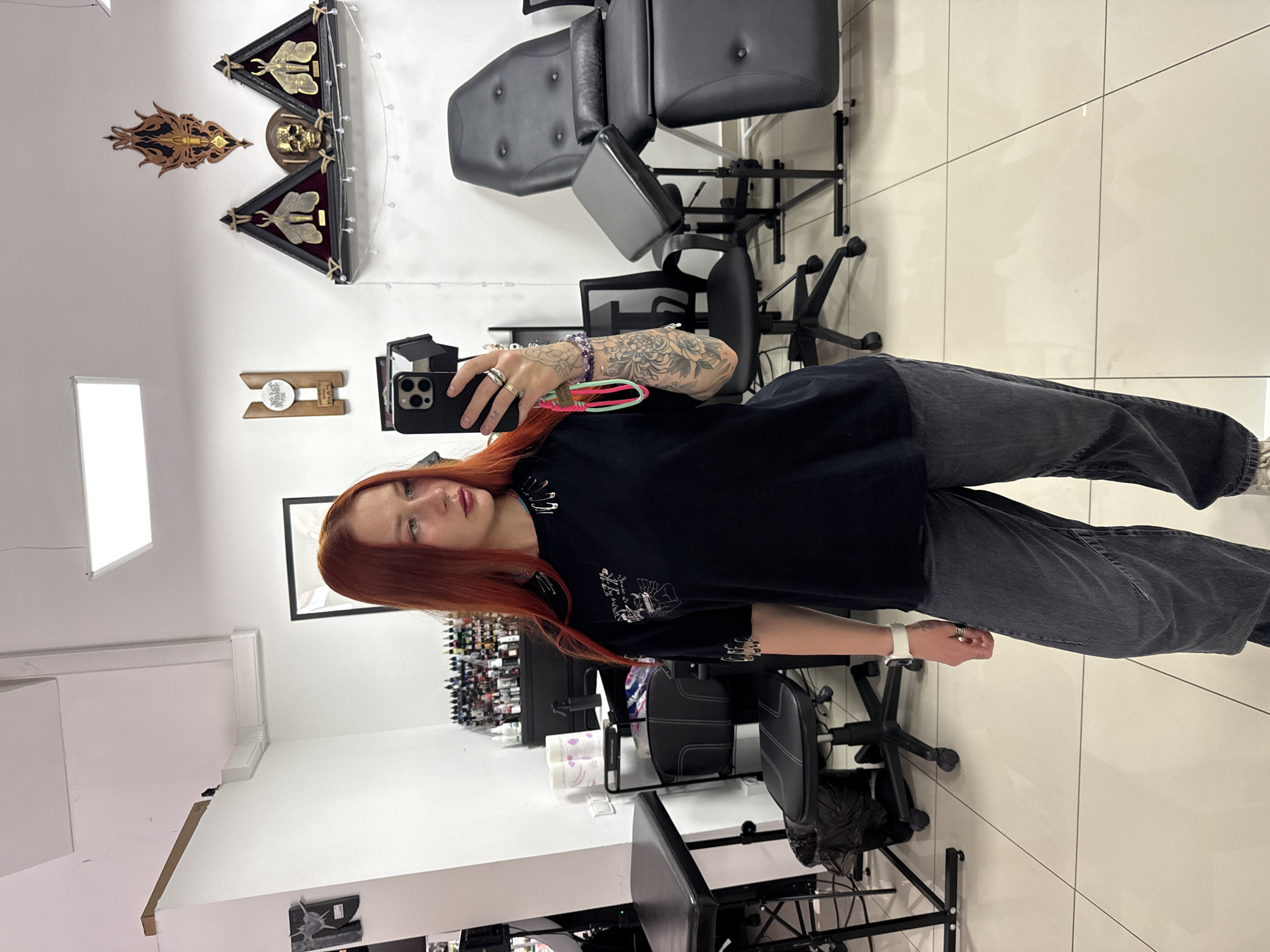 A woman with red hair is taking a selfie in a room. She is wearing a black t-shirt and gray jeans. The room contains tattoo equipment and chairs. The location is a tattoo studio, possibly in Rostov-on-Don, Russia.