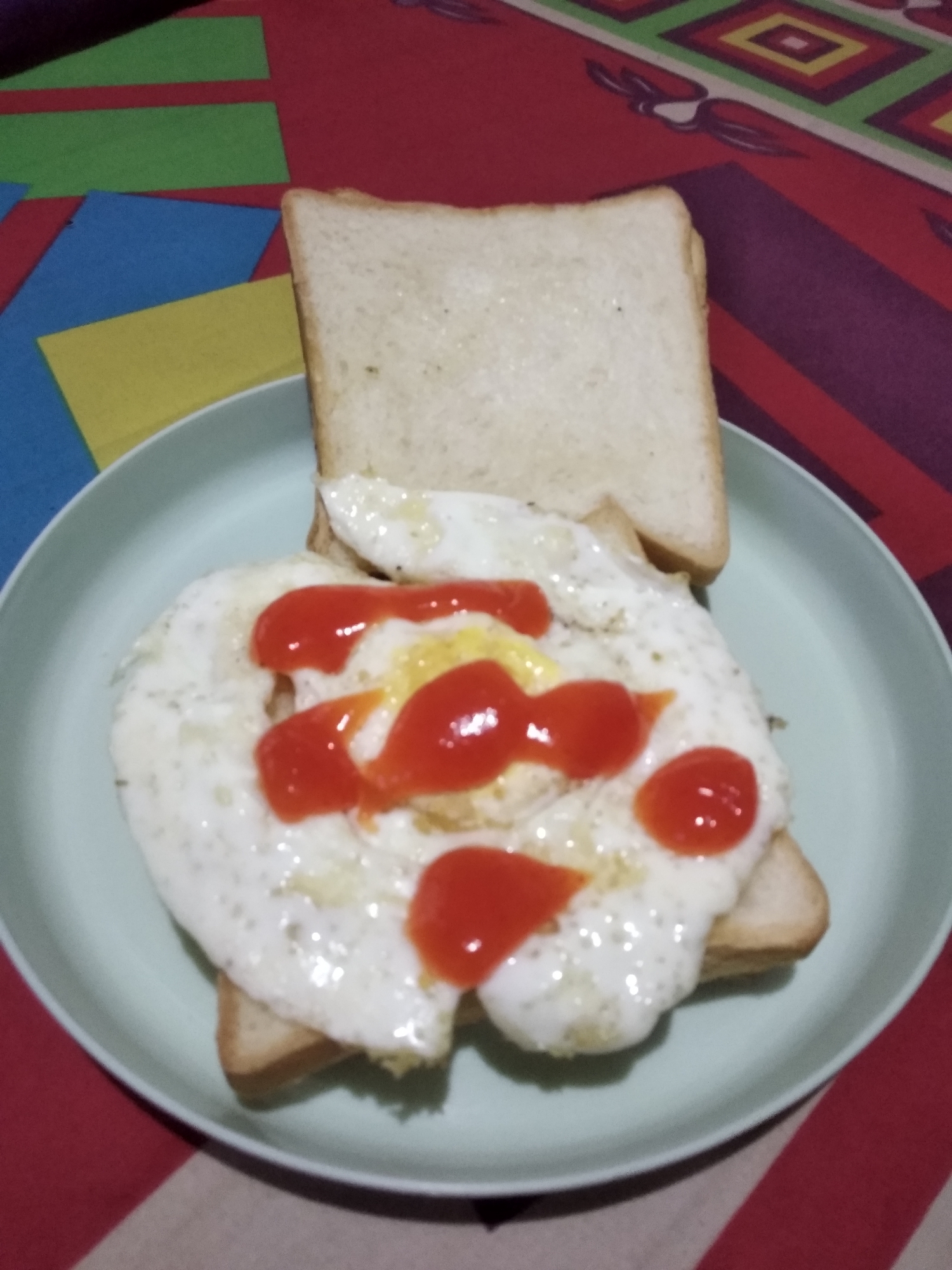 A close-up shot shows a plate of toast with a fried egg and ketchup. The toast is lightly browned and appears to be a whole wheat bread. The fried egg is cooked sunny-side up, with a slightly runny yolk. Ketchup is drizzled over the egg in several dollops. The plate is a pale green, and it rests on a colorful, patterned tablecloth. In the background, there are pieces of colorful paper, possibly for crafting or decoration. This image was likely taken indoors, in a residential setting in Balaraja, Indonesia, possibly on a table as part of a meal.