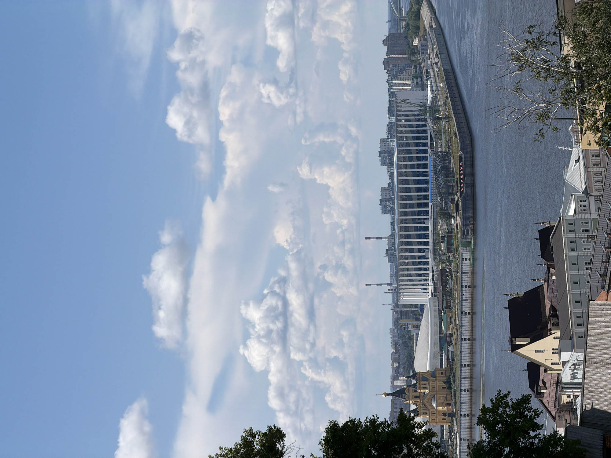 This is a landscape image of the city of Nizhny Novgorod, Russia, taken from a high vantage point. The view encompasses a wide river, a prominent stadium, a church, and various buildings. The sky is blue with scattered white clouds, suggesting a clear day.
In the foreground, there are trees on the left and right sides of the frame, partially obscuring the view. Below the trees, on the lower right, a cluster of multi-story buildings with dark-colored roofs is visible. These buildings appear to be a mix of residential and commercial properties, with some showing signs of age and others appearing more modern.
The middle ground is dominated by a wide, dark blue river, likely the Volga River, which is a major waterway in Russia. Along the riverbank, there is a promenade or embankment. On this embankment, to the left, stands a striking yellow church with dark domes and spires, characteristic of Russian Orthodox architecture. Further along the embankment, a large, modern stadium with a white facade and a distinctive seating structure is visible. The stadium has a sleek, contemporary design and appears to be a significant landmark.
In the background, the cityscape of Nizhny Novgorod extends towards the horizon. Numerous buildings of varying heights and architectural styles can be seen, including taller modern structures and older, lower buildings. Industrial elements, such as smokestacks, are also present in the distance. The overall impression is of a bustling city situated along a major river, with a blend of historical and modern architecture. The scene is serene and picturesque, with the natural beauty of the river and sky complementing the urban landscape.