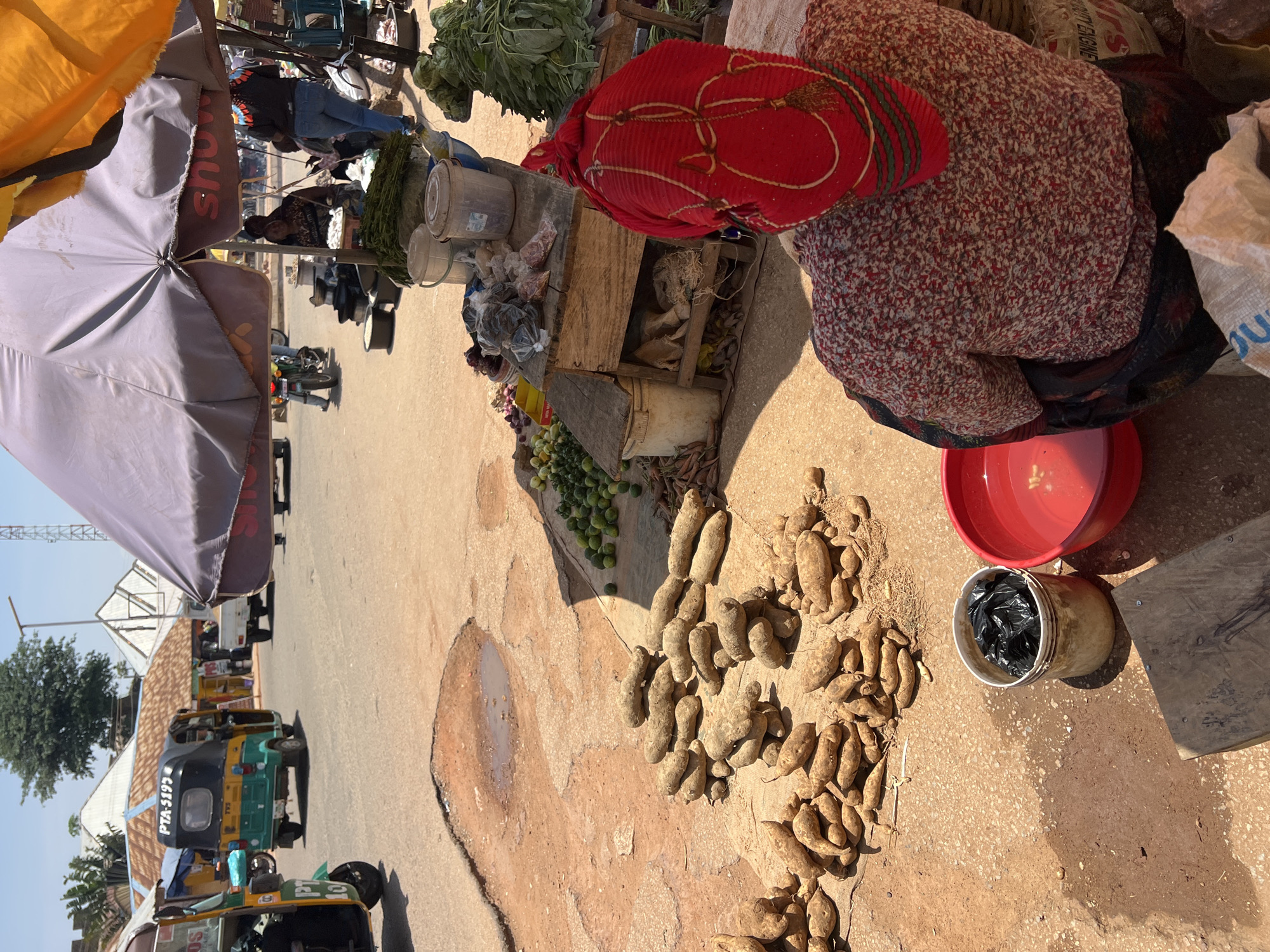 The image depicts an outdoor market scene in Jos, Nigeria, with a focus on a woman wearing a red headscarf and a floral-patterned shirt, seated and facing away from the camera. She is in the foreground, surrounded by various goods. To her left, a large red plastic basin filled with water is visible, next to a beige bucket containing black plastic bags. Piles of yams are arranged on the dusty ground in the mid-ground. Behind the woman, a wooden stall displays produce, including leafy green vegetables and what appear to be small gourds or limes. In the background, a busy street scene unfolds with a yellow and green auto-rickshaw, other vehicles, and market stalls under umbrellas, indicating a bustling commercial area. The ground is a mix of dry dirt and patchy puddles, characteristic of the environment in Jos.