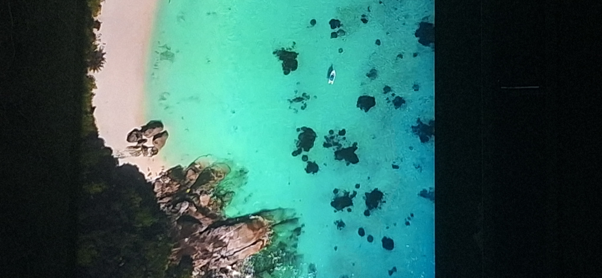 An aerial view shows a tropical island coastline with lush green vegetation bordering a white sandy beach. The water is a clear turquoise, with dark rocks protruding from the sea in various locations. A small white boat is visible in the water. The image does not depict the city of Bila Tserkva, Ukraine.