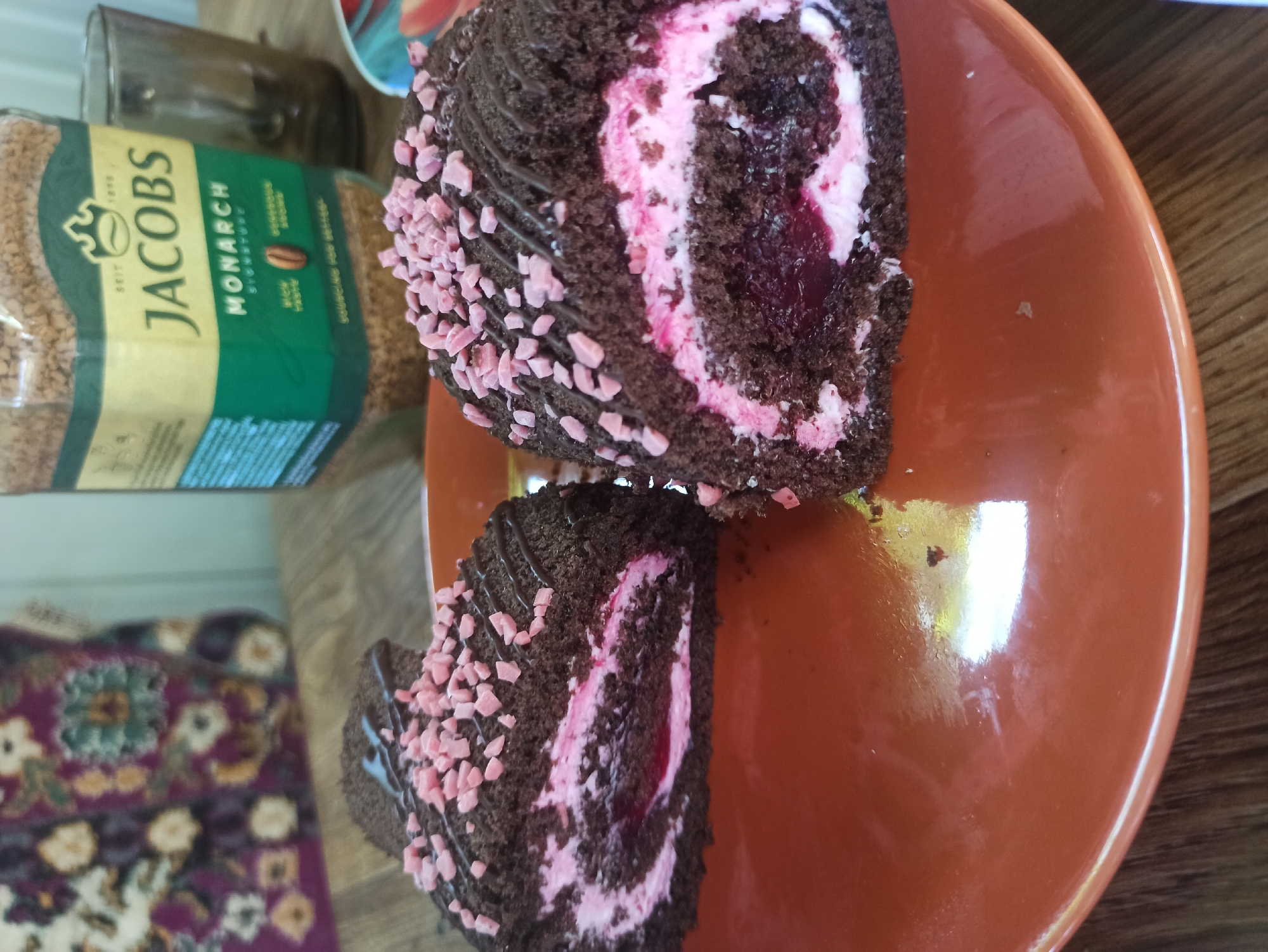 Two slices of chocolate Swiss roll cake sit on an orange ceramic plate. The cake has a dark chocolate exterior, a pink cream filling, and a dark red cherry or berry filling in the center. The top of the cake is drizzled with chocolate and sprinkled with small pink pieces, likely candy. In the background, a jar of Jacobs Monarch instant coffee is visible, along with a glass with some dark liquid. A colorful fabric with floral patterns is also in the background. The scene appears to be set on a wooden surface. The image was taken in Kryvyi Rih, Ukraine.