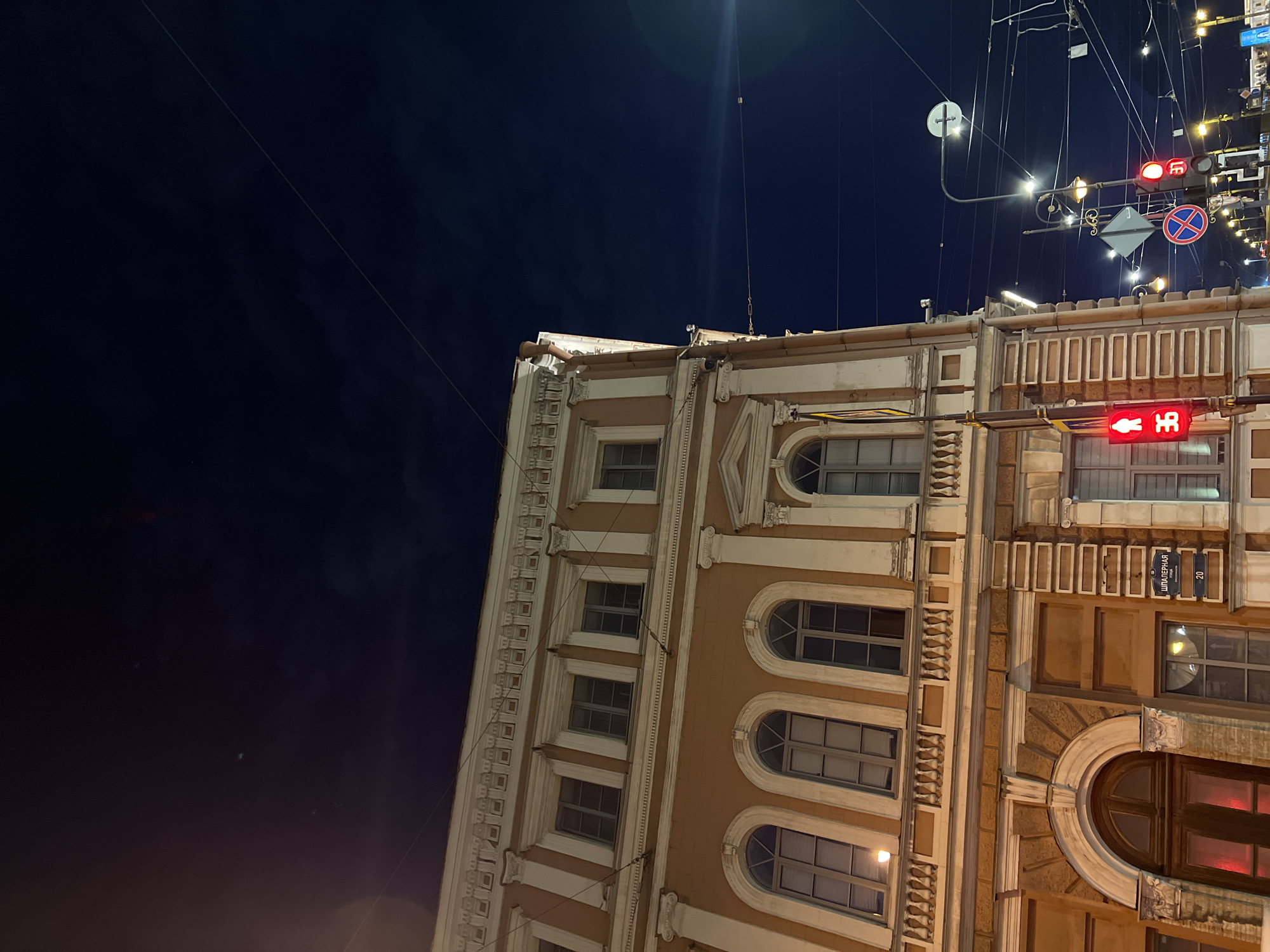 The image shows a building in Saint Petersburg, Russia, at night. The building is multi-story with an ornate facade, including windows with arched tops. Street signs are visible, including a red traffic light indicating "stop." Utility lines and streetlights are also visible in the dark sky. The image is taken from a low angle, looking up at the building. The building has a sign that reads "Шпалерная улица 20," indicating its location on Shpalernaya Street, number 20.