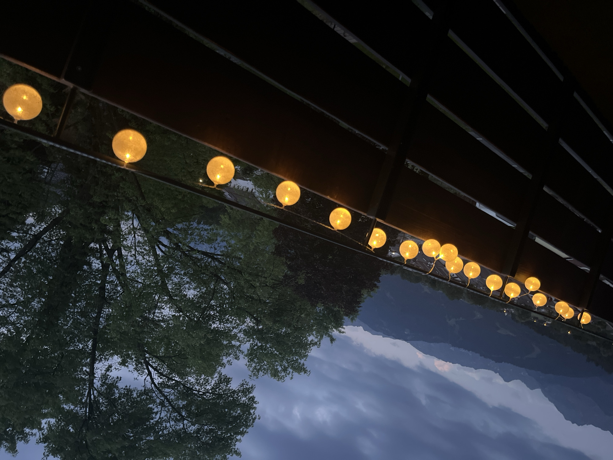 The image depicts an outdoor scene at dusk, likely a balcony or patio overlooking a mountain range. String lights with spherical, yellow bulbs are strung along a dark brown wooden railing, casting a warm glow. The lights are illuminated, indicating it is nighttime or twilight.
In the background, a dramatic mountain landscape is visible under a cloudy, dark blue sky. The mountains appear to be steep and covered in dark vegetation. Faint lights from buildings or settlements can be seen on the slopes of the mountains.
To the right of the frame, lush green trees with dense foliage dominate the midground and extend into the background. The overall atmosphere is serene and moody, with the soft light from the string lights contrasting with the darkening sky and the imposing mountains.
The location is identified as Garmisch-Partenkirchen, Germany, a Bavarian town known for its alpine scenery. This setting is consistent with the visual elements in the image. There are no people or animals visible in the frame.