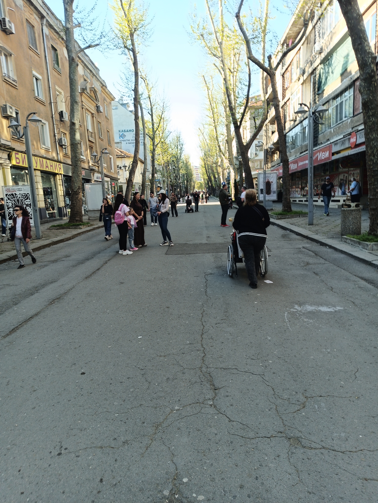 The image depicts a street in Burgas, Bulgaria. It's a paved road lined with bare trees and buildings on either side. The sky is clear and bright.
Several people are present. On the left side of the road, there's a woman walking and a group of people gathered. Further down the street, there are more individuals walking or standing. On the right side of the street, a person is pushing someone in a wheelchair.
The buildings lining the street appear to be a mix of residential and commercial properties, with shops and businesses visible at street level. The road itself shows signs of wear and tear, with cracks and uneven surfaces. The scene suggests a pedestrian-friendly area, with people strolling and interacting in the street.