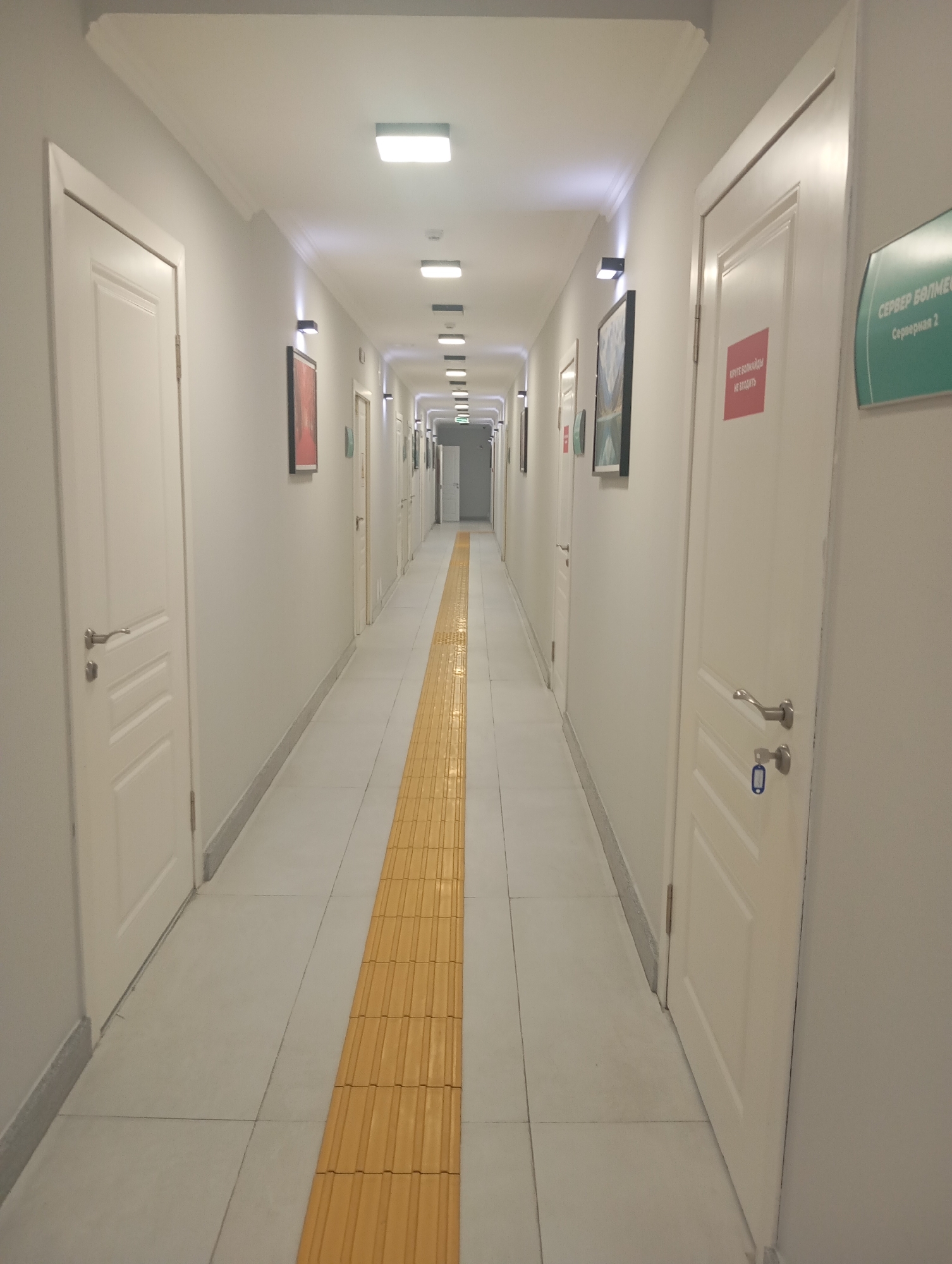 The image shows a long, brightly lit hallway in what appears to be a modern office building or institution. The hallway is lined with identical white doors on both sides, with the exception of the one on the right which has a sign above it. The floor is tiled in a light grey color, and a textured yellow tactile paving runs down the center of the hallway, indicating a pathway for visually impaired individuals. Several square, modern light fixtures are mounted on the ceiling, providing uniform illumination.
On the walls, between the doors, are framed pictures and some have small black square lights above them, suggesting they are art installations or decorative elements. One door on the right side of the hallway has a green sign with white text that reads "СЕРВЕР БОЛМЕСІ" which translates to "SERVER ROOM" in Kazakh, followed by "Серверная 2" which translates to "Server Room 2". There is also a red rectangular sign on one of the doors that reads "ЎРГЕ БОЛМАЙДЫ" which translates to "DO NOT ENTER" in Kazakh.
The hallway extends into the distance, creating a sense of depth and perspective. The overall impression is one of orderliness and functionality. The architecture suggests a relatively new construction.
While the image depicts a generic hallway, the presence of Kazakh language on the signs strongly suggests that this location is in Kazakhstan. Almaty, being the largest city in Kazakhstan, is a plausible location for such a building. However, without further context or identifying landmarks, pinpointing the exact location within Almaty is not possible from the image alone.