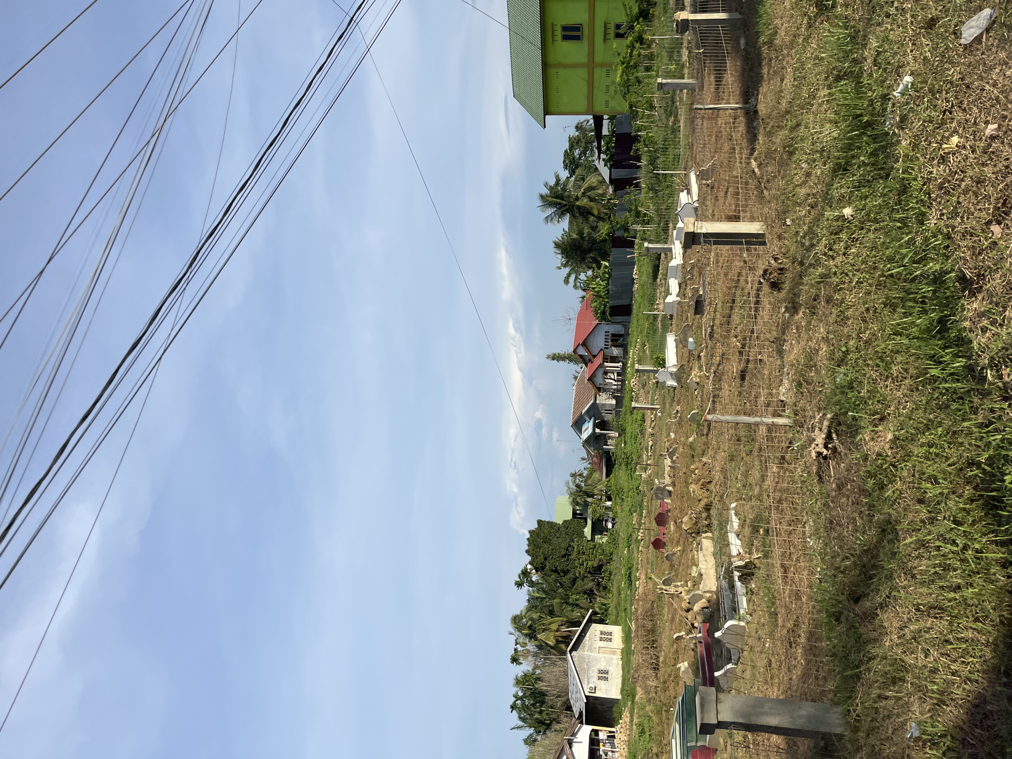 This is an eye-level, outdoor, medium shot of a cemetery in Landuh, Indonesia. The cemetery is in the foreground, with tombstones scattered across dry, patchy grass. A wire fence encloses part of the cemetery, with concrete posts. In the midground, there are several houses with red and green roofs, as well as lush green trees. A bright green, two-story building is prominent on the right side of the image. The background shows a clear blue sky with some scattered white clouds. Utility wires are visible in the upper portion of the image, stretching across the sky. There are no people or animals visible in the scene. The overall impression is one of a rural or semi-rural community.