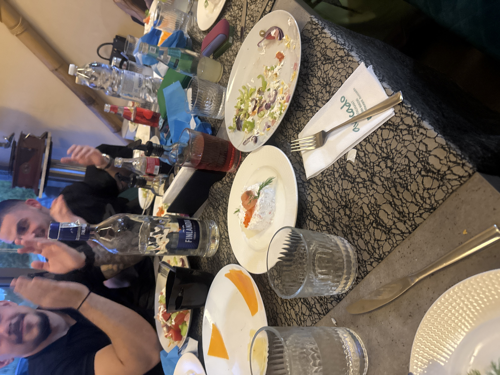 The image captures a scene on a tabletop covered with a grey patterned surface. Numerous plates, some with leftover food remnants, are visible. A half-empty Finlandia vodka bottle is present, along with several glasses. One glass contains a red liquid, likely wine. Cutlery, including a fork on a napkin bearing the "Avocado" restaurant logo, is scattered around. The background reveals two men, one closer to the camera with dark hair and a beard. It is impossible to determine the exact location in Plovdiv, Bulgaria, without further information.