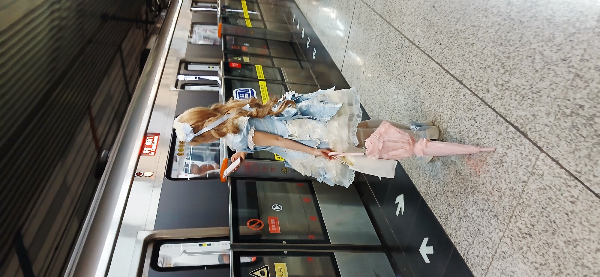 A woman in a blue and white dress with blonde hair is standing on a subway platform in Chongqing High-tech Zone, China. She is holding a pink umbrella and a mobile phone, and she is standing in front of a subway train. The platform is made of gray tiles.