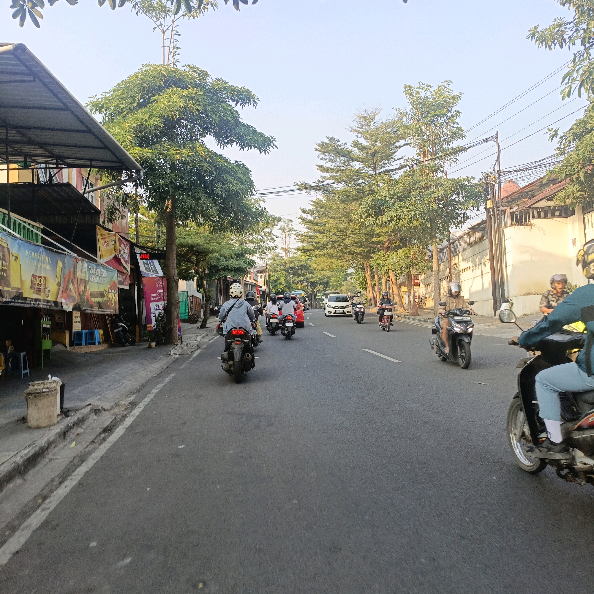 The image depicts a street scene in Salatiga, Indonesia, likely in the morning or late afternoon given the sunlight. The street is paved and appears to be a two-way road with a white line marking the center. On the left side of the road, there is a sidewalk with several shops and buildings. Banners and signs, some in Indonesian, are visible on the shopfronts. A few blue plastic stools are placed outside one of the establishments. A large, cylindrical concrete trash receptacle is also present on the sidewalk. Trees with dense green foliage line both sides of the street, creating a shaded environment.
The street is active with motorcycle traffic. Several individuals are riding motorcycles, some with passengers. Most riders are wearing helmets. One person on a motorcycle in the foreground is wearing a teal-colored jacket and light blue pants. Another person on a motorcycle further down the road is wearing a tan jacket and shorts. A white car is also visible in the middle of the traffic. The buildings on the right side of the street are mostly residential, with tiled roofs and white or beige walls. Overhead, power lines are strung between poles. The sky is clear and light blue.